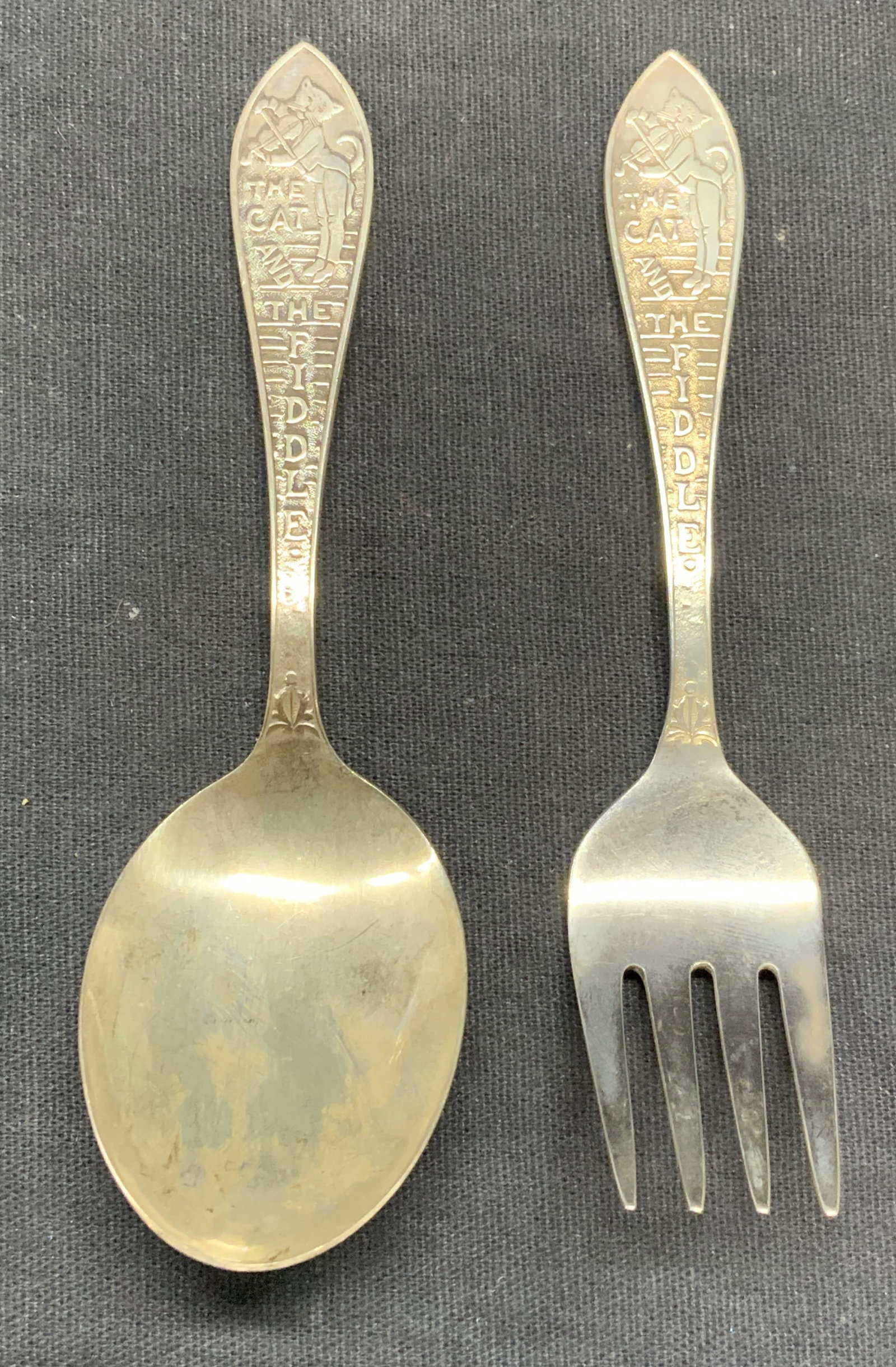 2 Vintage Cat & Fiddle Sterling Silver Utensils: Pair of matching Cat and The Fiddle motif sterling silver childrens fork and spoon. Marked sterling to underside, larger piece measures approx. 4.25 inches long. Total weight of both pieces .87 ozt. B