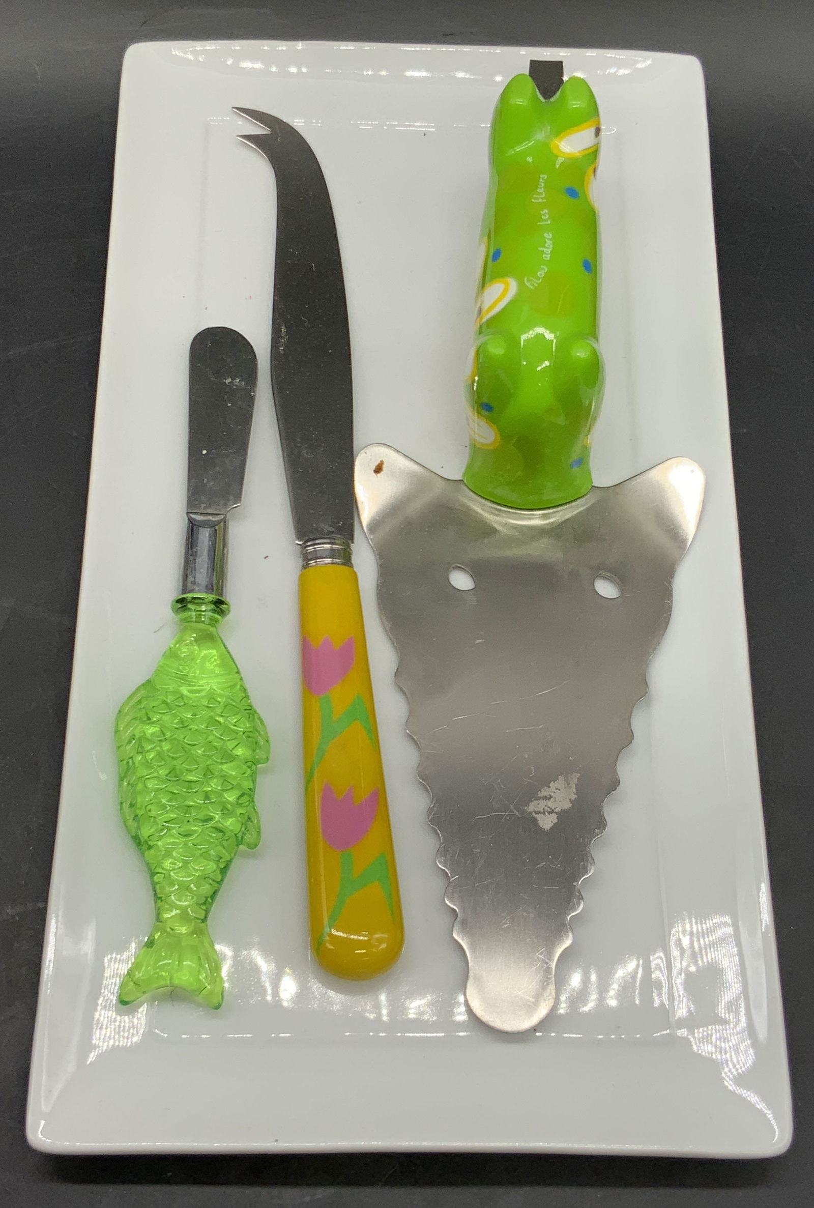Lot 4 Serving Plate & Utensils, Kitchenware: Three serving utensils and one white rectangular ceramic serving plate. The plate measures approx. 6.25L x 11.75W in. Serving utensils feature bright, colorful handles. Brands include Palate & Plate,