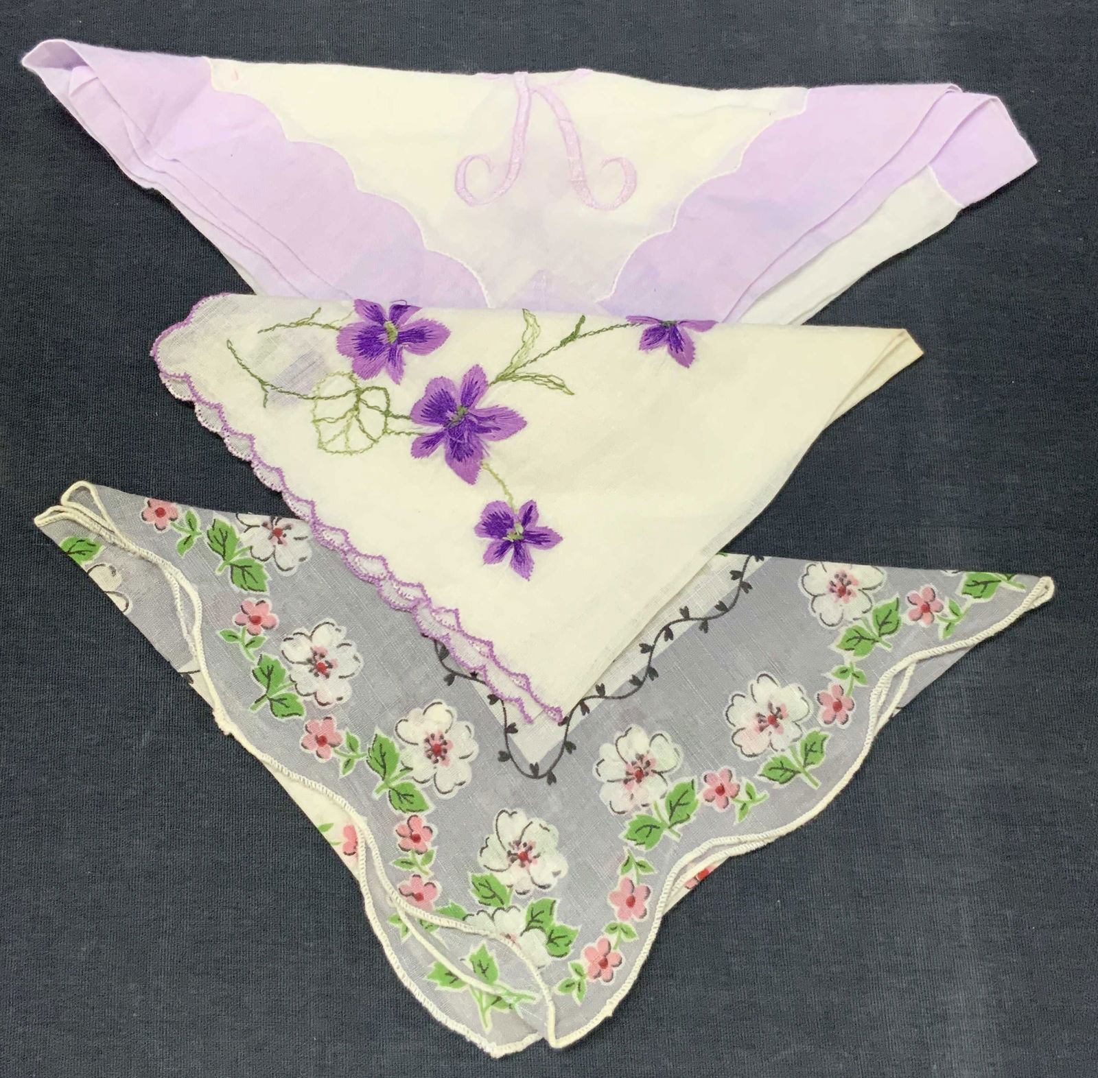 Lot 3 Vtg Embroidered Floral Linens: Lot of 3 vintage linens. 1 with purple embroidered K with purple edges, 1 with dark purple embroidered flowers with scalloped edges and 1 handkerchief with printed white and pink flowers on gray and w