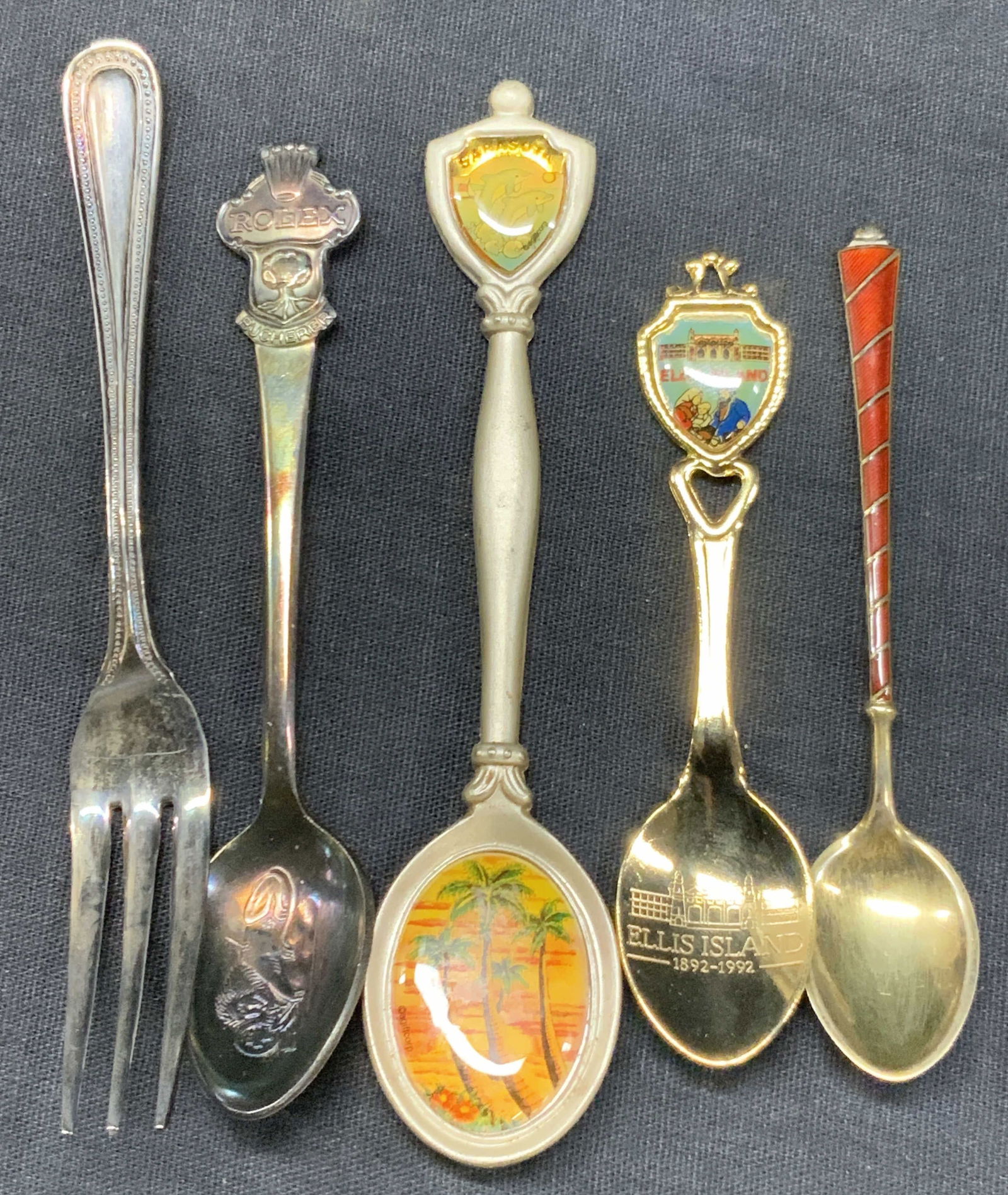 Lot 5 Vtg Sterling Silver & Pewter Utensils: Lot of 5 vintage utensils, 1 marked steering silver with red enamel handle, 1 Godinger fork, 1 souvenir spoon from Ellis island in gold tones. 1 pewter spoon from Sarasota and 1 spoon marked Rolex, Sw