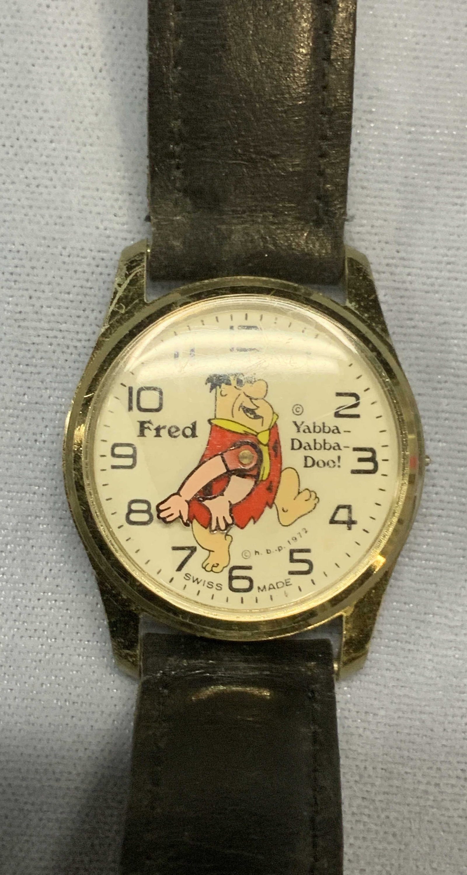 1972 HBP Fred Flintstone Gold Tone Watch: Vintage 1972 HBP Fred Flintstone gold tone Watch with black leather strap. Watch face measures approx. 1.25 inches wide. Not tested for function. Some surface wear including scratching observed. Needs