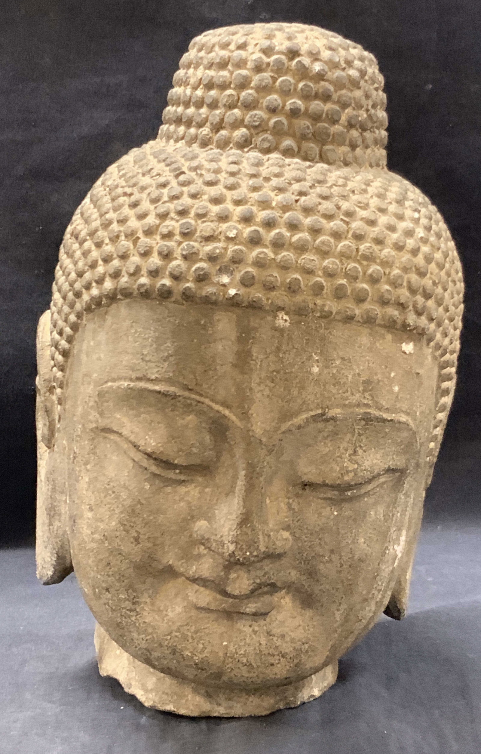 Carved Stone Buddha Head: Buddha head made of carved stone with top knot, long ear lobs and eyes closed in mediation. Approx 10.5 inches tall and 5 x 6 inches. Evident weight from stone and moderated surface wearing due to age