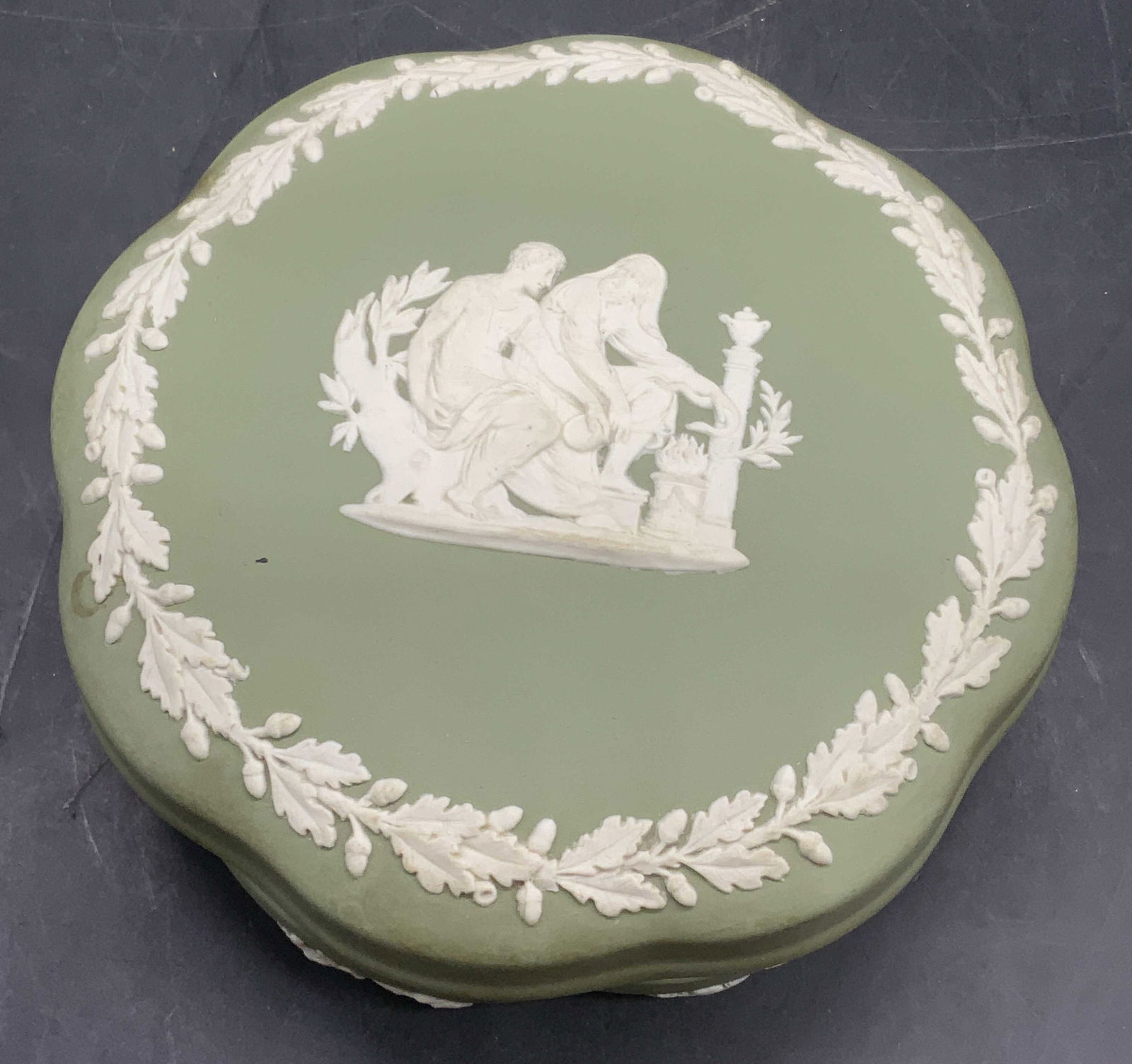 WEDGWOOD Vintage Green Jasperware Trinket Box, Eng: A vintage green Jasperware lidded trinket box featuring a scalloped rim. Greco Roman motif. Raised white figurals, putti, laurels, and more throughout. Measures approx. 1.75H x 4.75W in. Underside mar