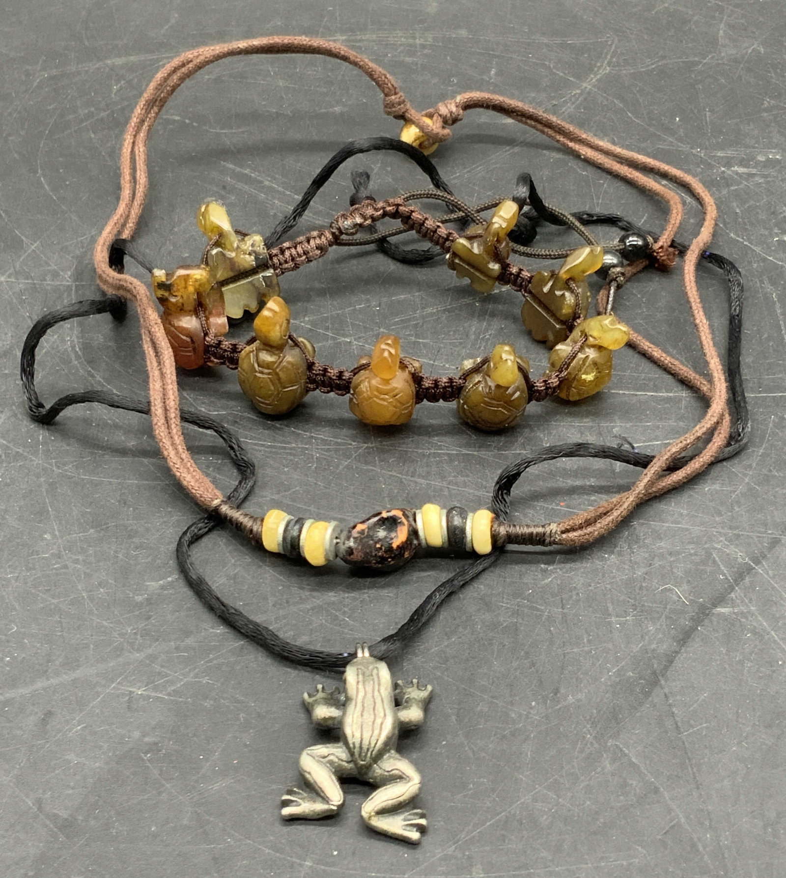 Lot 3 Cord Necklaces & Bracelet, Jewelry: Three pieces of cord / string jewelry featuring natural stones and metal beads. One necklace features a silver tone metal frog figural pendant and the other necklace features stone, metal, and composi