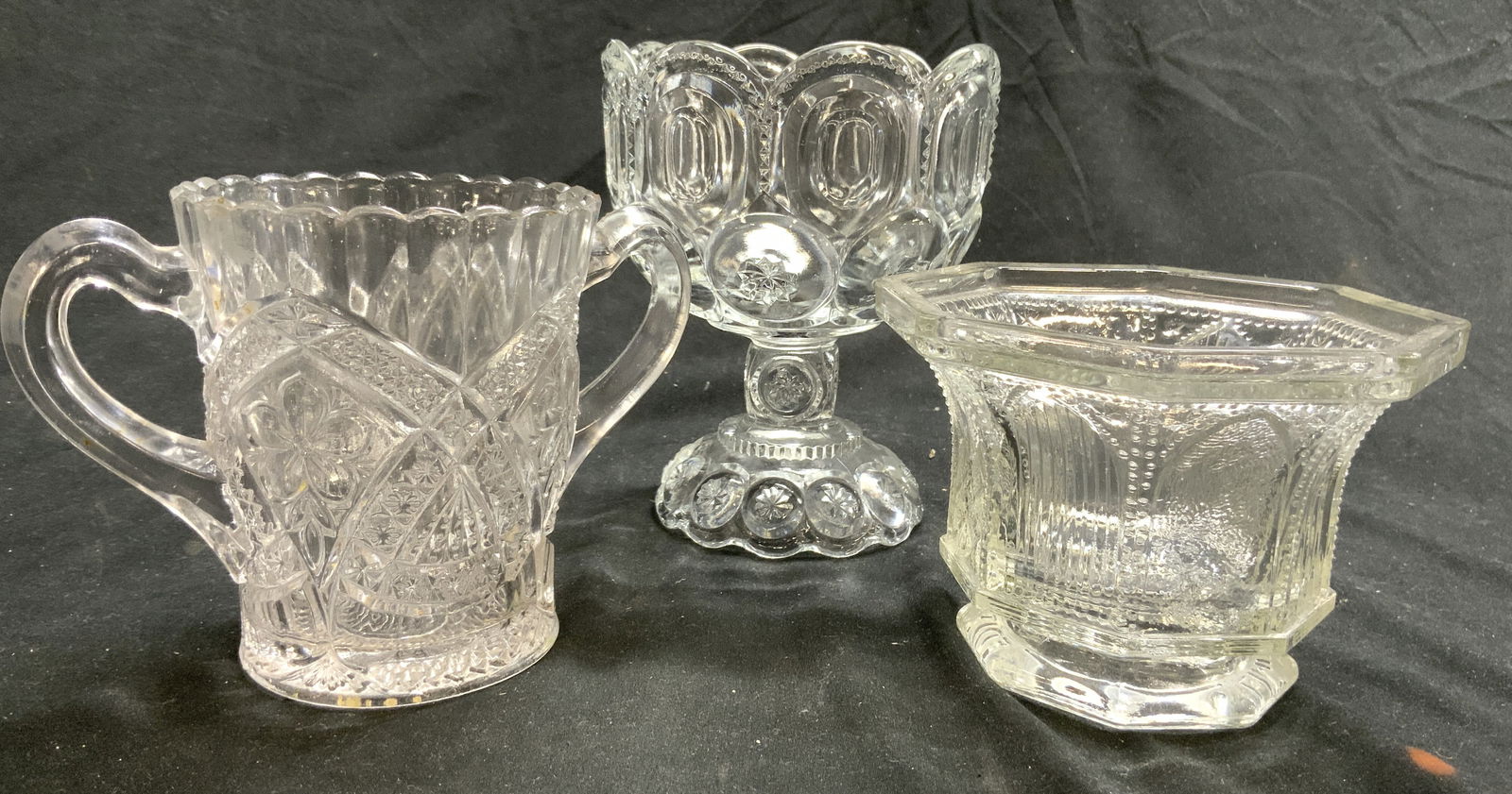 Lot 3 Assorted Art Glass Vessels: Lot includes 1 pedestaled goblet style candy dish with large curved tooth rim oval and other geometric pattern detail, 1 double handle vessel with geometric and floral relief detail, & 1 footed vessel