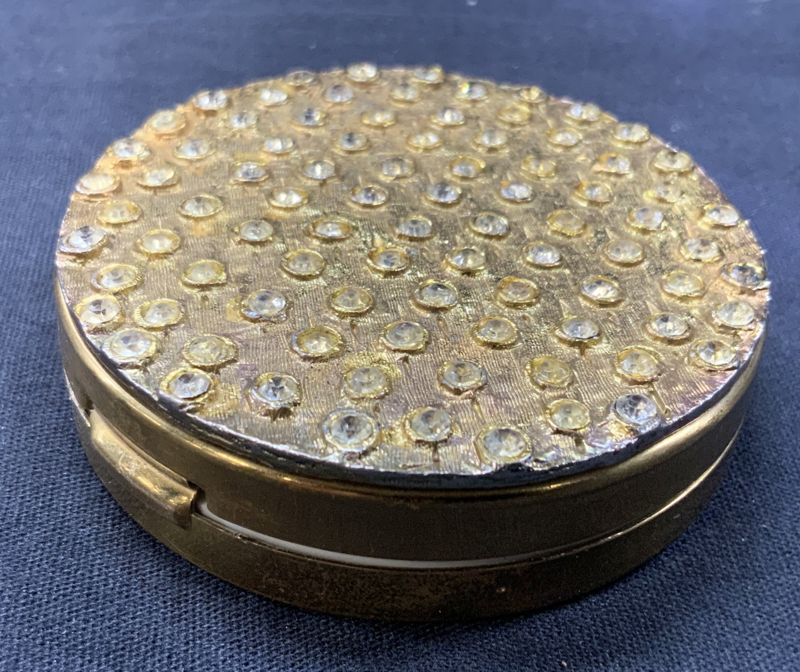 Vntg Brass Jeweled Blush Compact: Vintage blush compact, made of brass or gilt metal with lid encrusted with clear crystals. Approx 3 inch diameter. Interior has original blush puff and inner mirror. Evident wearing to metal and crack