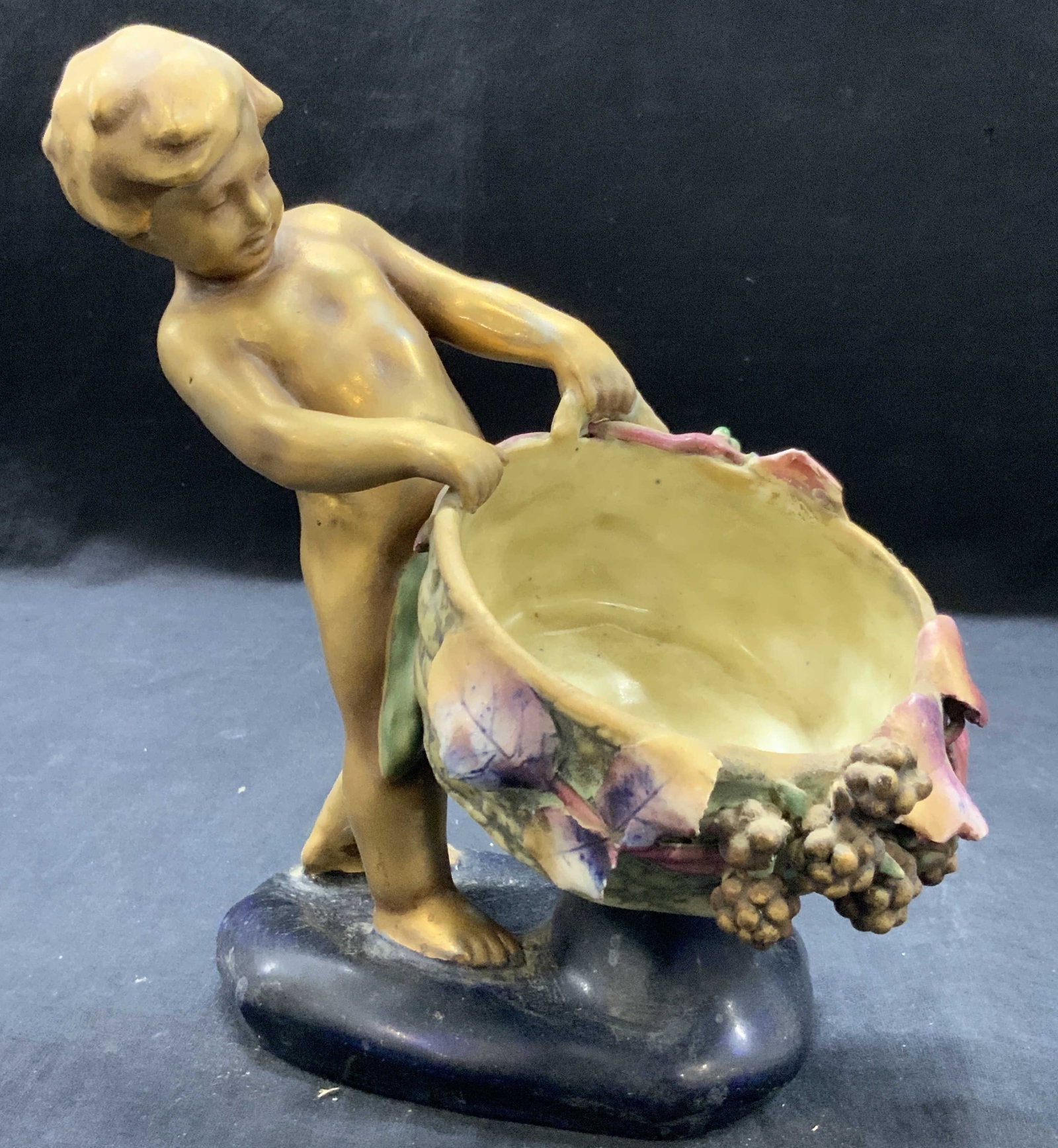 Stamped Gilt Porcelain Cherub Figural: Stamped on underside, with a crown motif. A porcelain figure of a gilt child holding a basket with flowers on the sides. Approx 10 inches tall and 8 inches long. Base being 5 x 4 inches Evident losses