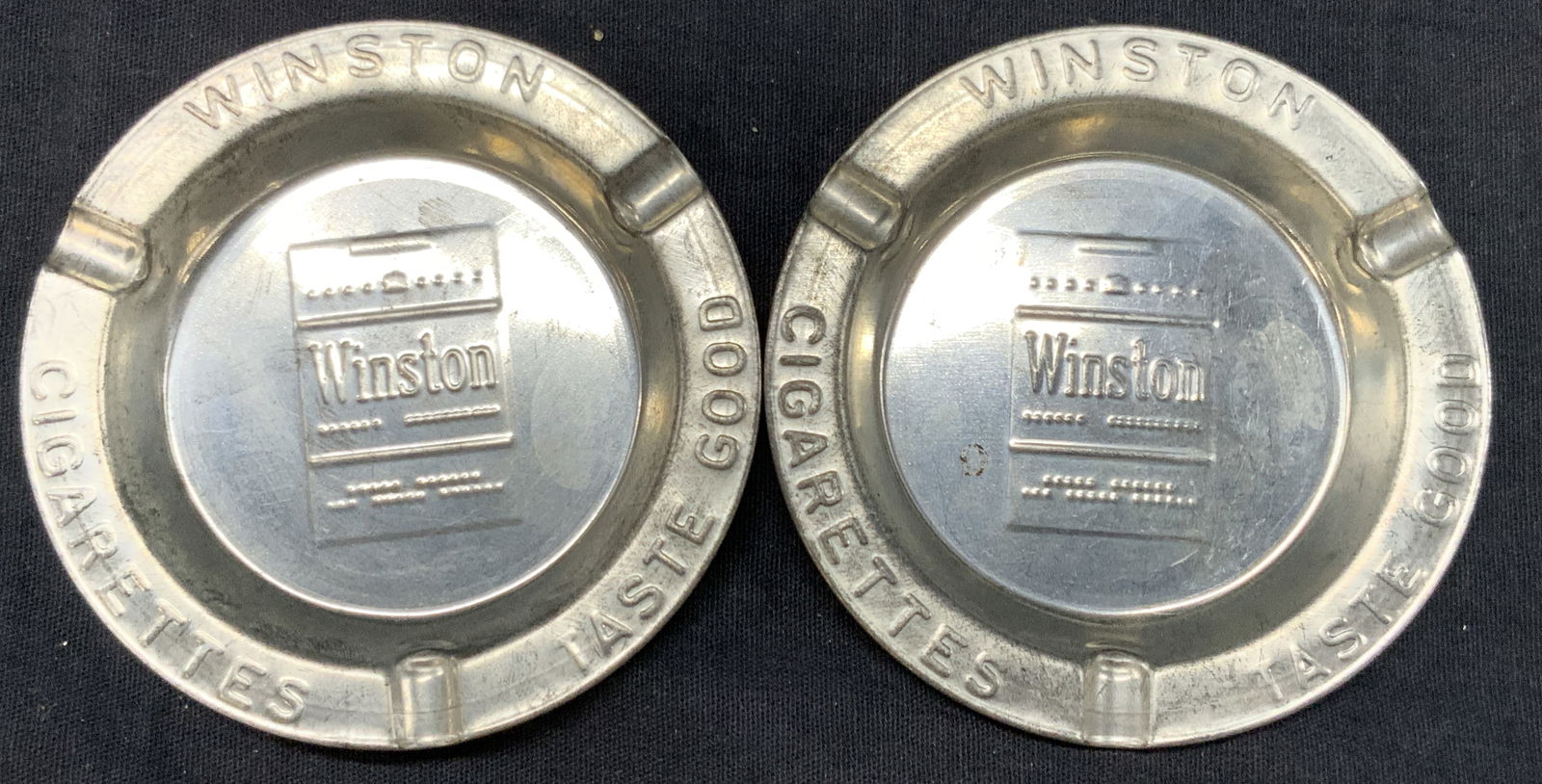 Pr Winston Cigarettes Aluminum Ash Trays: Pair of vintage aluminum ash trays. Embossed and reading Winston cigarettes, Taste good with a motif of the cigarette packs on the center. Approx 3.25 inch diameter, Moderate wearing to metal due to a