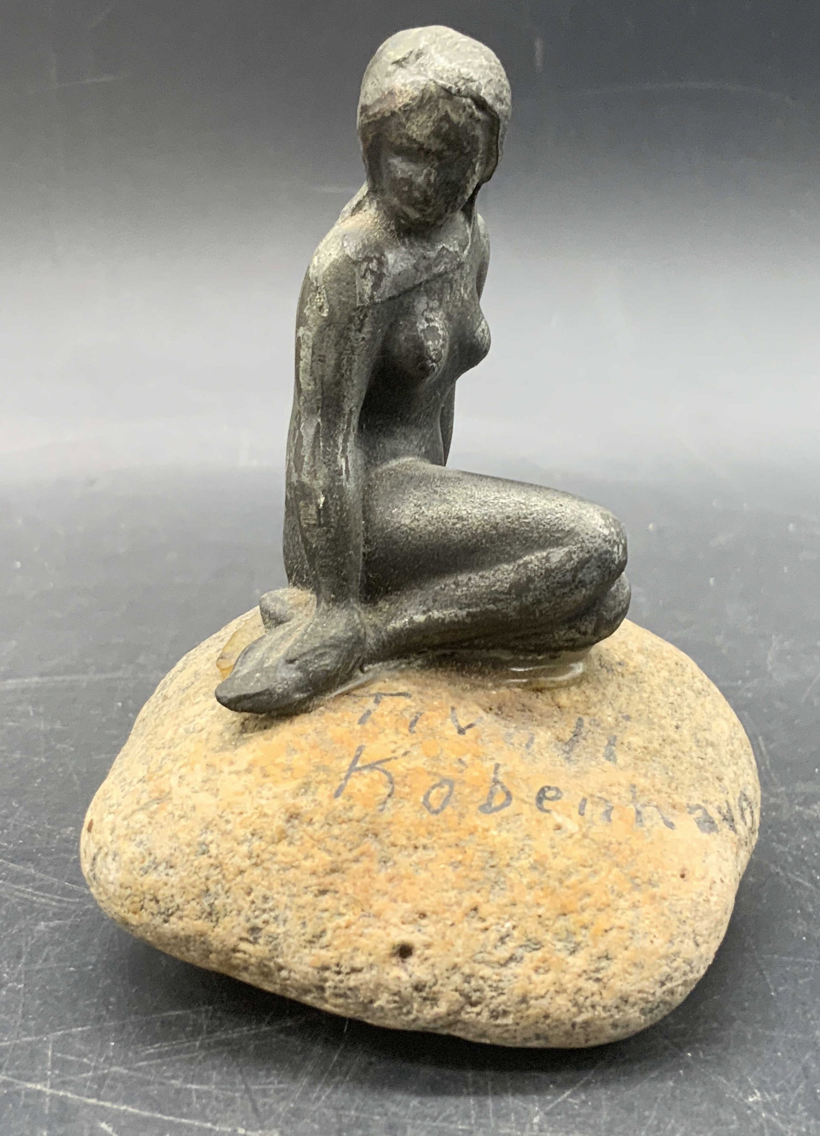 TIVOLI KOBENHAVN Metal Mermaid On Stone Figural: A metal mermaid figural seated atop a stone. Obverse reads TIVOLI KOBENHAVN. Mermaid is affixed to rock. Measures approx. 3.75H x 2.75W in. Some surface wear and residue present. Collectibles, home de