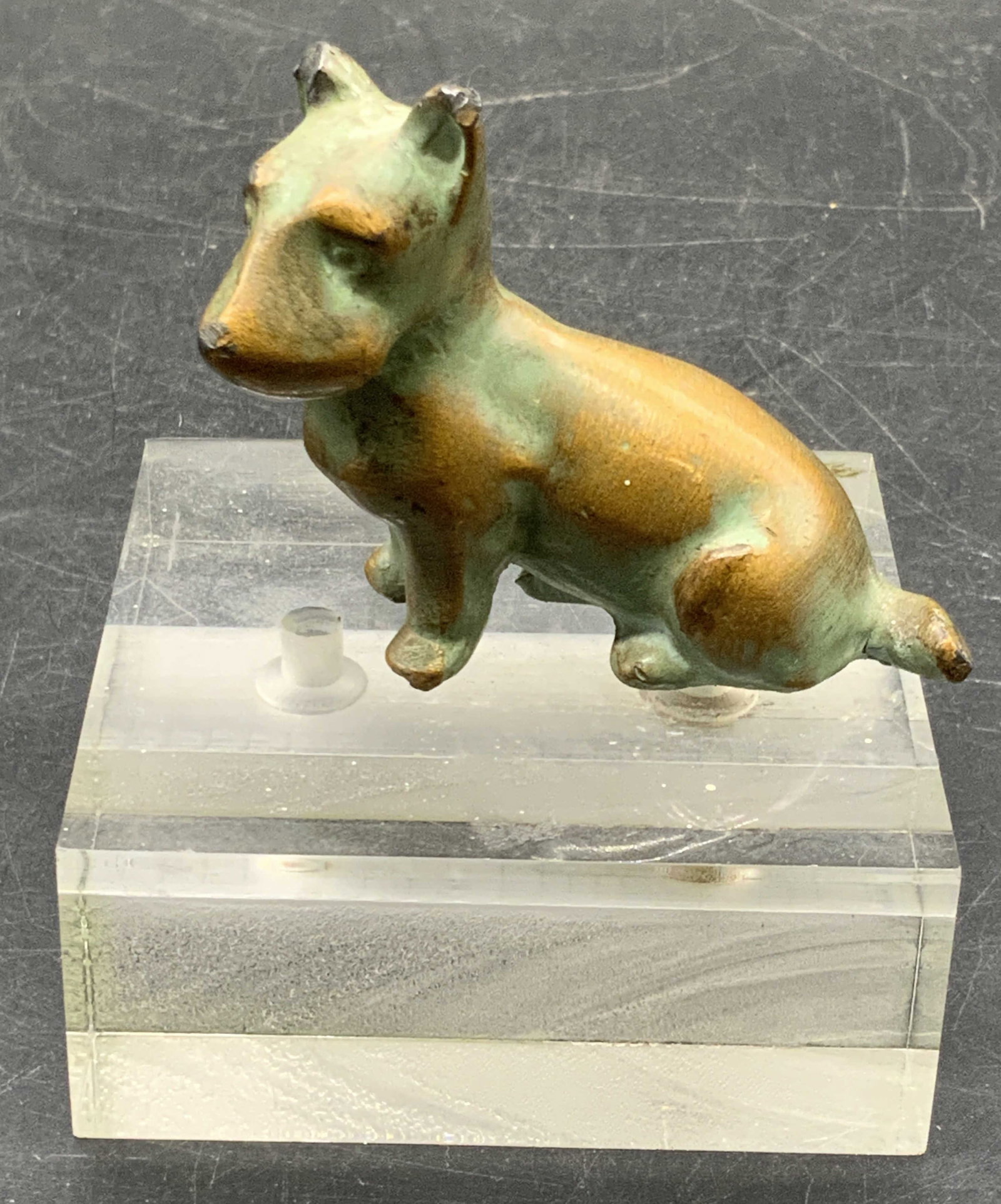 Metal Dog Figural W/ Chunky Acrylic Base: A metal dog figural attached to a clear chunky acrylic base. The dog is crafted from metal and has a green & bronze hued finish. Pieces are connected via a screw to the underside. The position of the 