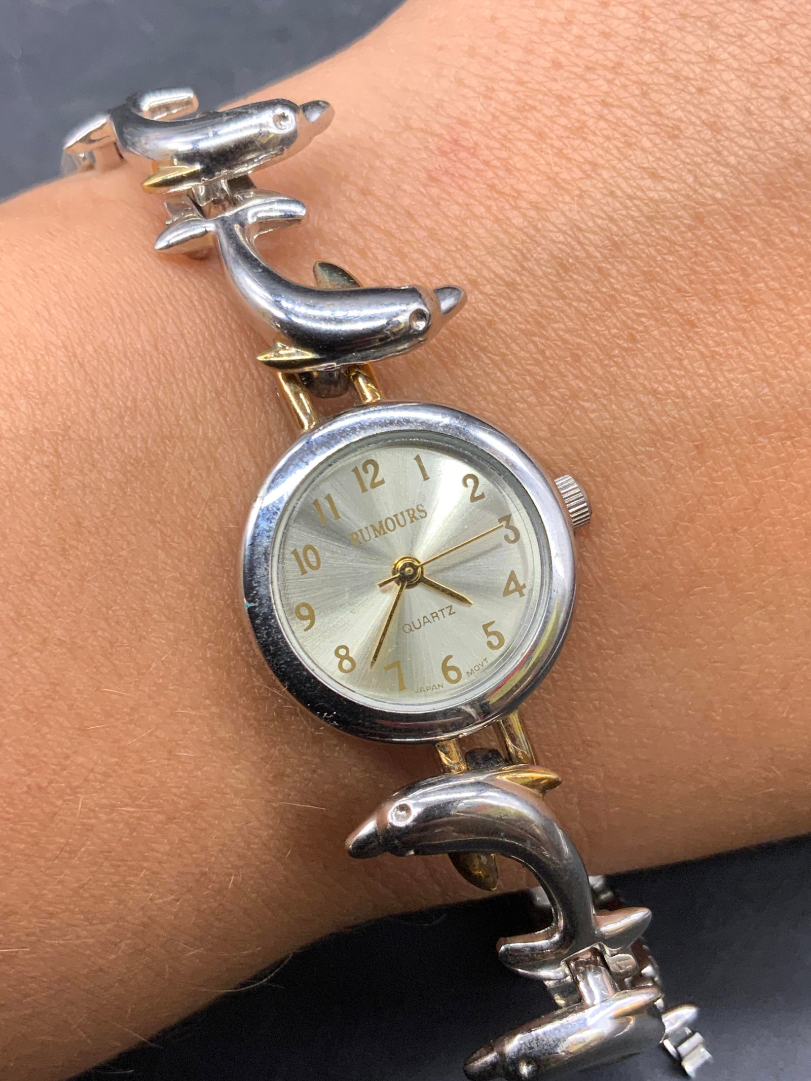 RUMOURS Quartz Dolphin Motif Wristwatch: A silver and gold tone wristwatch featuring a circular watch face. Secures with clasp closure. Watch face reads RUMOURS QUARTZ. Reverse reads Stainless Steel Back, 48593. Watch length measured end to 