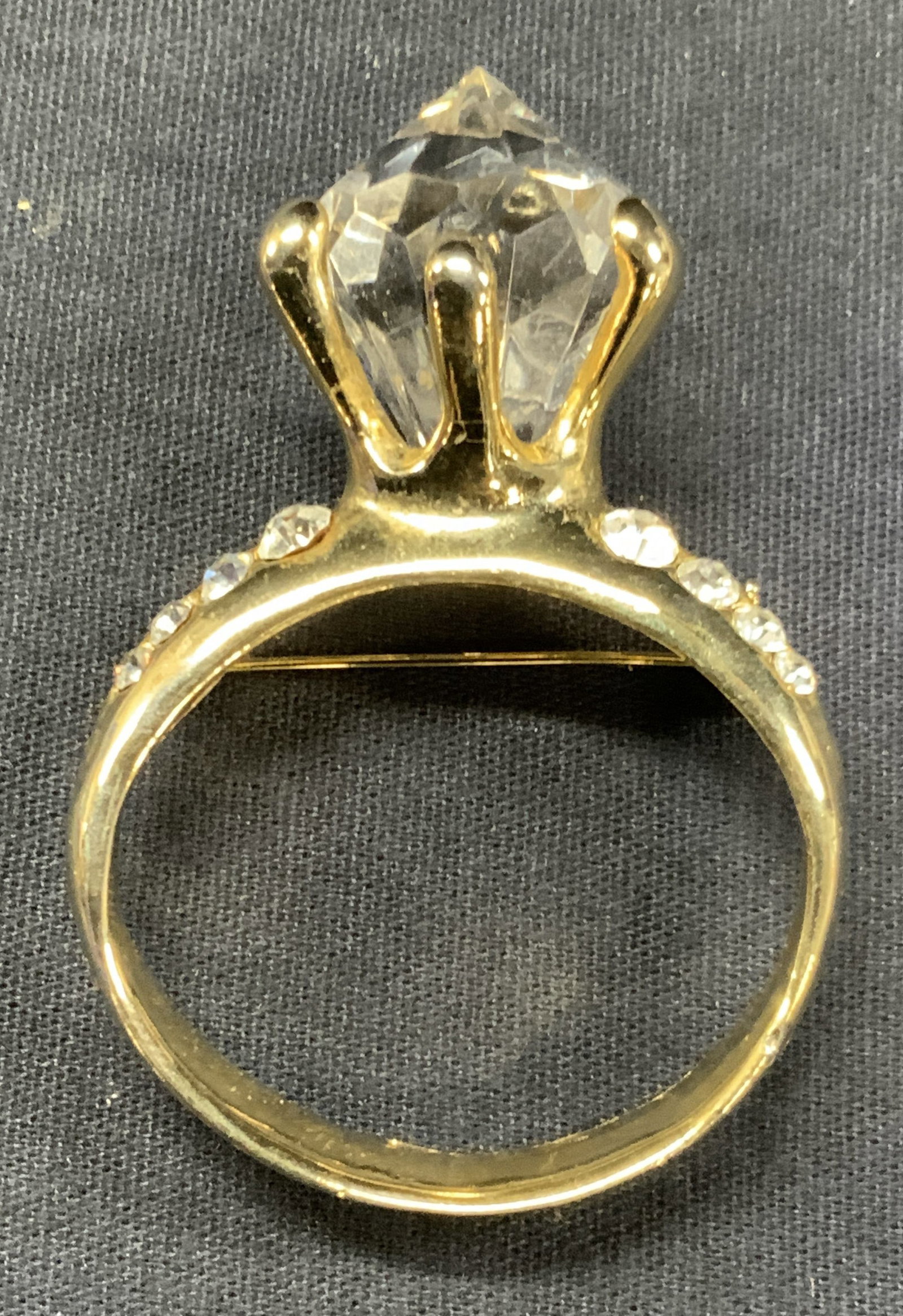 Vntg AJC Crystal Engagement Ring Brooch: Vintage AJC engagement ring brooch made of gold toned metal with faceted crystal center stone and flanked by encrusted crystals. Approx 3 x 2 inches Moderate surface wearing, scratches observed. Brooc