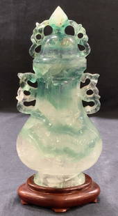 Carved Stone Asian Vessel W Stand: Carved Quartz stone, clear, green and purple tones throughout with intricate carved handles and topper. Comes with carved wooden base of red wood Approx 10 inches tall and 4 x 2.5 inches wide.