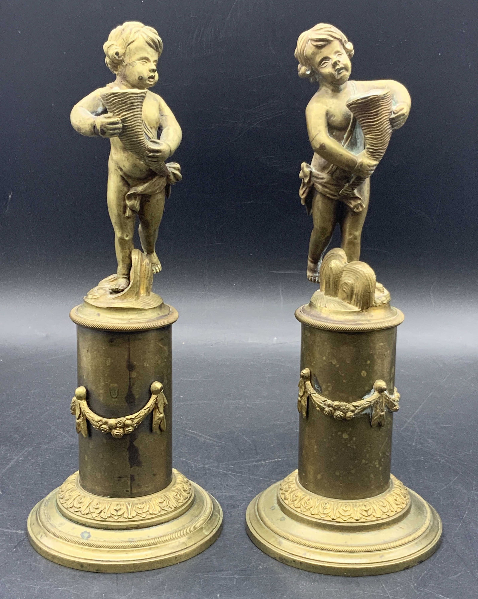 Pair of Gilt Brass Cherub Figurals: Pair of Gilt Brass Cherub Figurals. Cherubs are holding a cornucopia and base is accented with an embossed neoclassical laurel. Each Figural measures 9.5 in height. The base on each figural measures 3