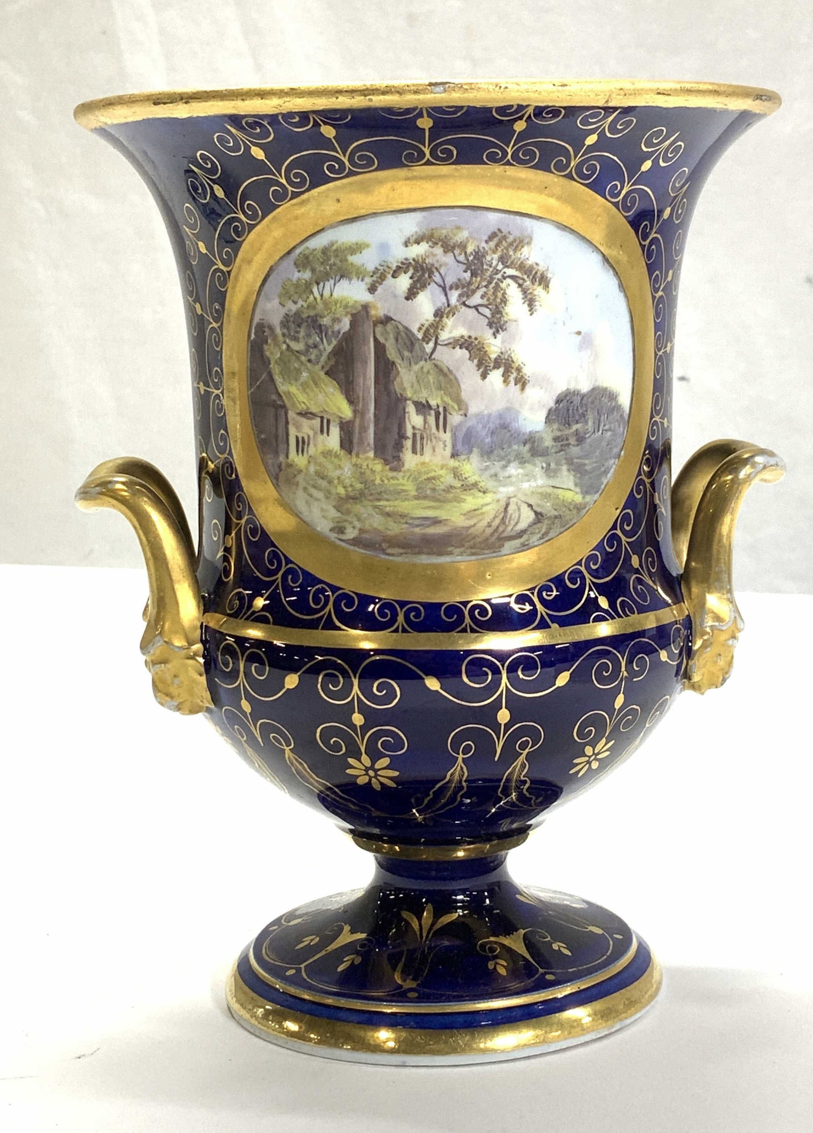 European Hand Painted Porcelain Urn with Gilt: Royal Crown Derby -inspired hand painted porcelain urn in cobalt with gilt scrolls, lion head Figurals at base of the handles, and painted thatched roof English cottages. No hallmarks. Measures 5.5 in