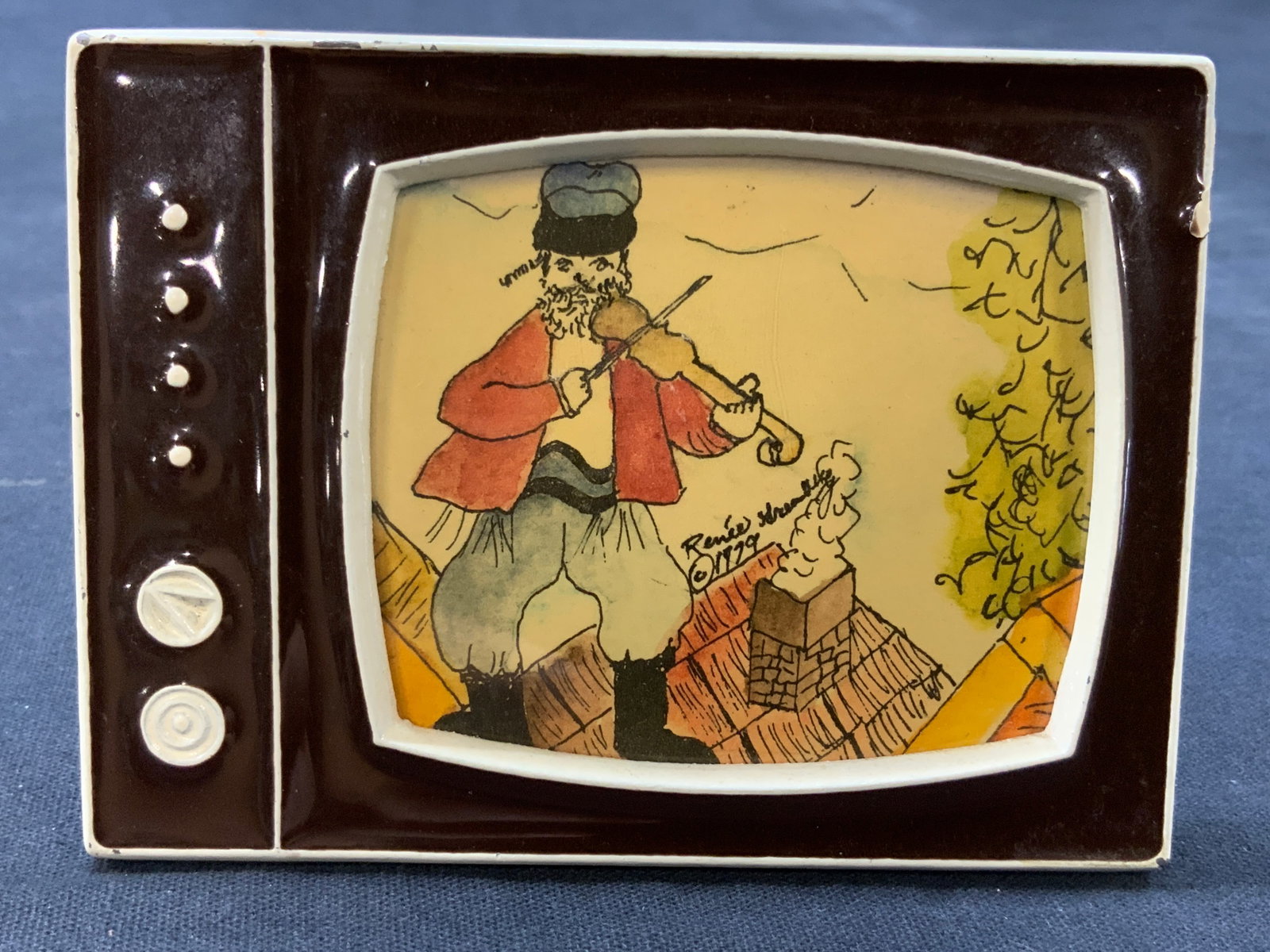 Sgd 1979 Fiddler On The Roof Watercolor: Signed on center, Possibly reads Renee, 1979. Depicts the fiddler playing his fiddle while standing on a roof. Framed in an enamel frame made to resemble a vintage TV. Approx 3 x 4 inches window measu