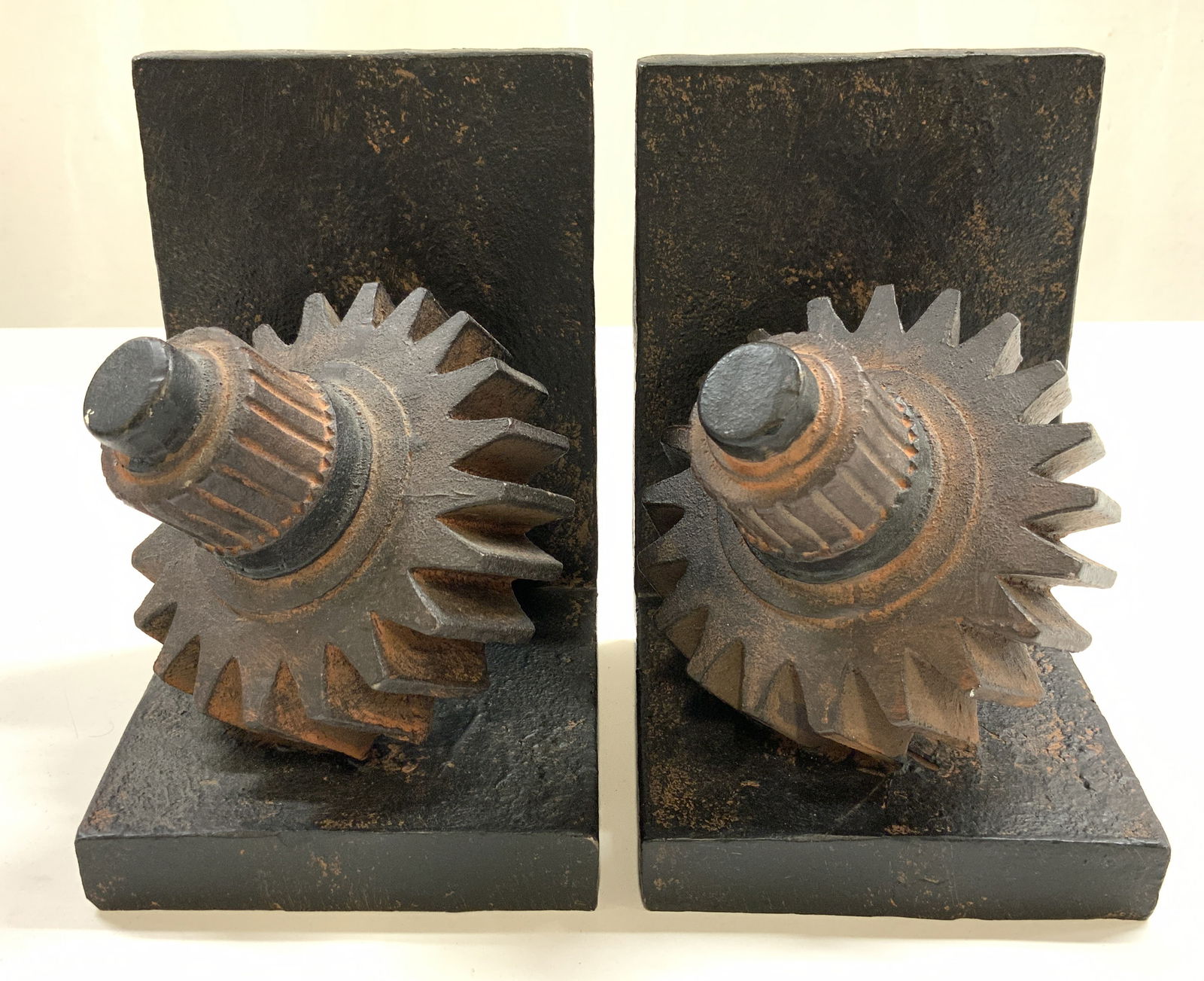 Pair Polystone Gear Bookends: Lot includes 2 bookends. Each appear to be Polystone. Each bookend has gear form. Each are black and rust toned. Bookends each measure approx 6.25 inches tall 4.25 inches wide 4.75 inches deep. Booken