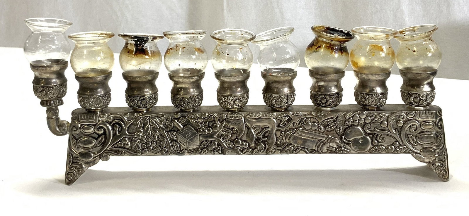 Silver Pl Embossed Menorah w 9 Glass Holders: Long menorah for table or hearth with embossed motifs of dreidel, torah scroll, wine grapes, and scroll designs. 9 glass candle holder cup inserts. Measures 11.5 in. Long, height 3.5 in. , 1.25 in. Wi