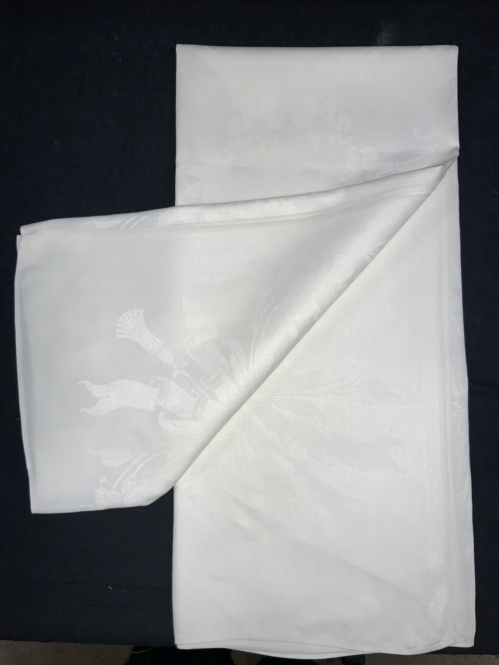 Vntg Cotton & Linen Lily Tablecloth: Vintage white toned linen and cotton blend tablecloth with imagery of Lily flowers on edges and ribbon motifs. Approx 108 x 50 inches Minor wearing to tablecloth due to age and use. Vintage tablecloth