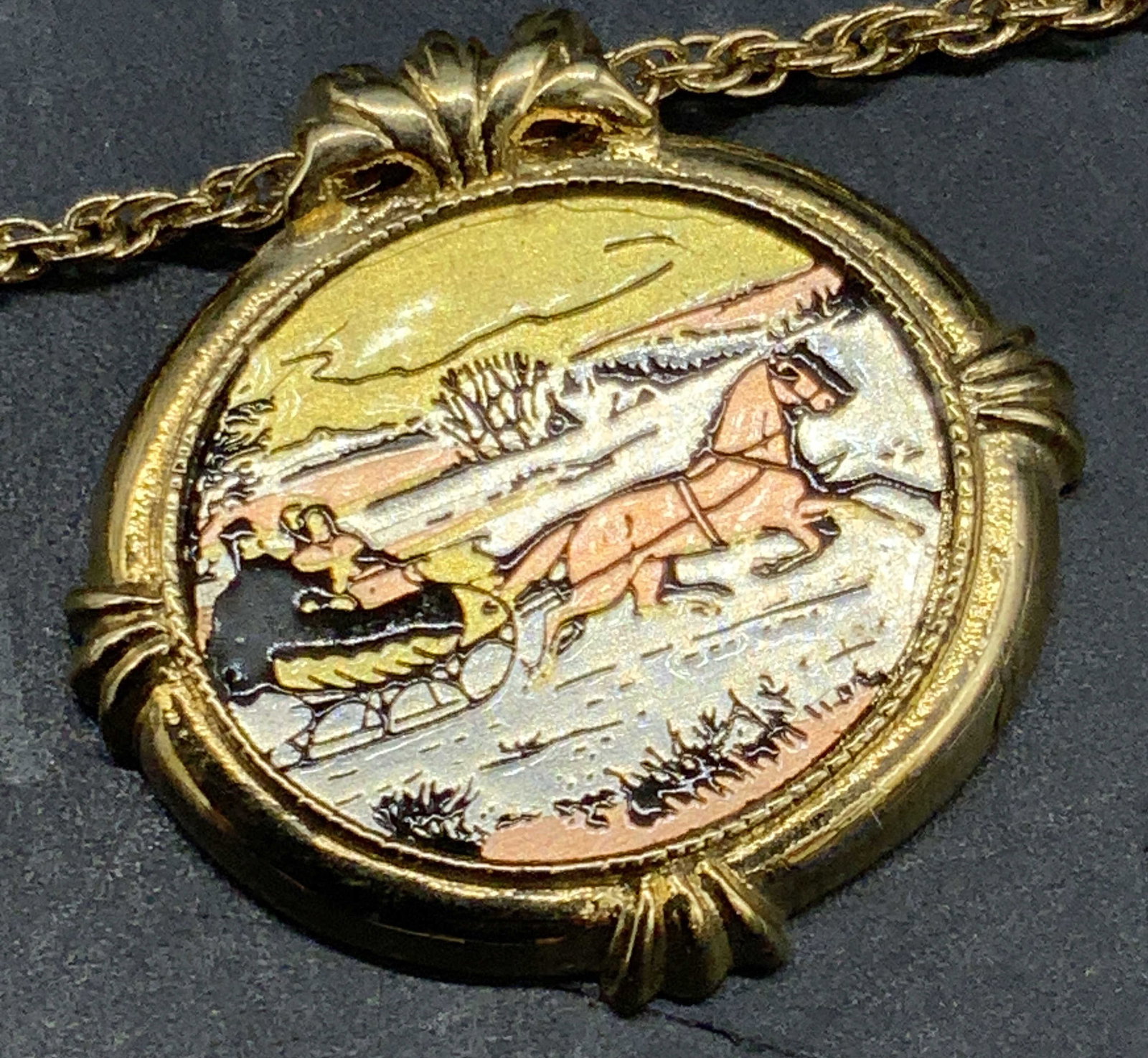 1982 AVON COUNTRY CHRISTMAS Pendant Necklace: A gold tone metal chain necklace featuring an oval form pendant. The obverse scene features a couple sitting in a sled that is pulled by a horse. The reverse reads 1982 AVON COUNTRY CHRISTMAS. Necklac