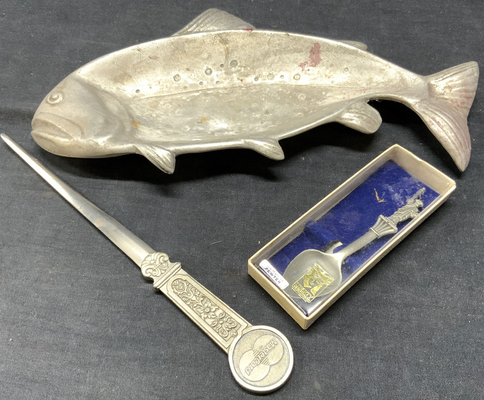 Lot 3 Pewter Tabletop Accessories: Lot includes 1 fish form serving dish (signed illegibly, 1 TUMASEK PEWTER MASTERCARD letter opener, & 1 LIBERTY condiment spoon with original box, tag reads GENUINE PEWTER. Fish form serving dish meas