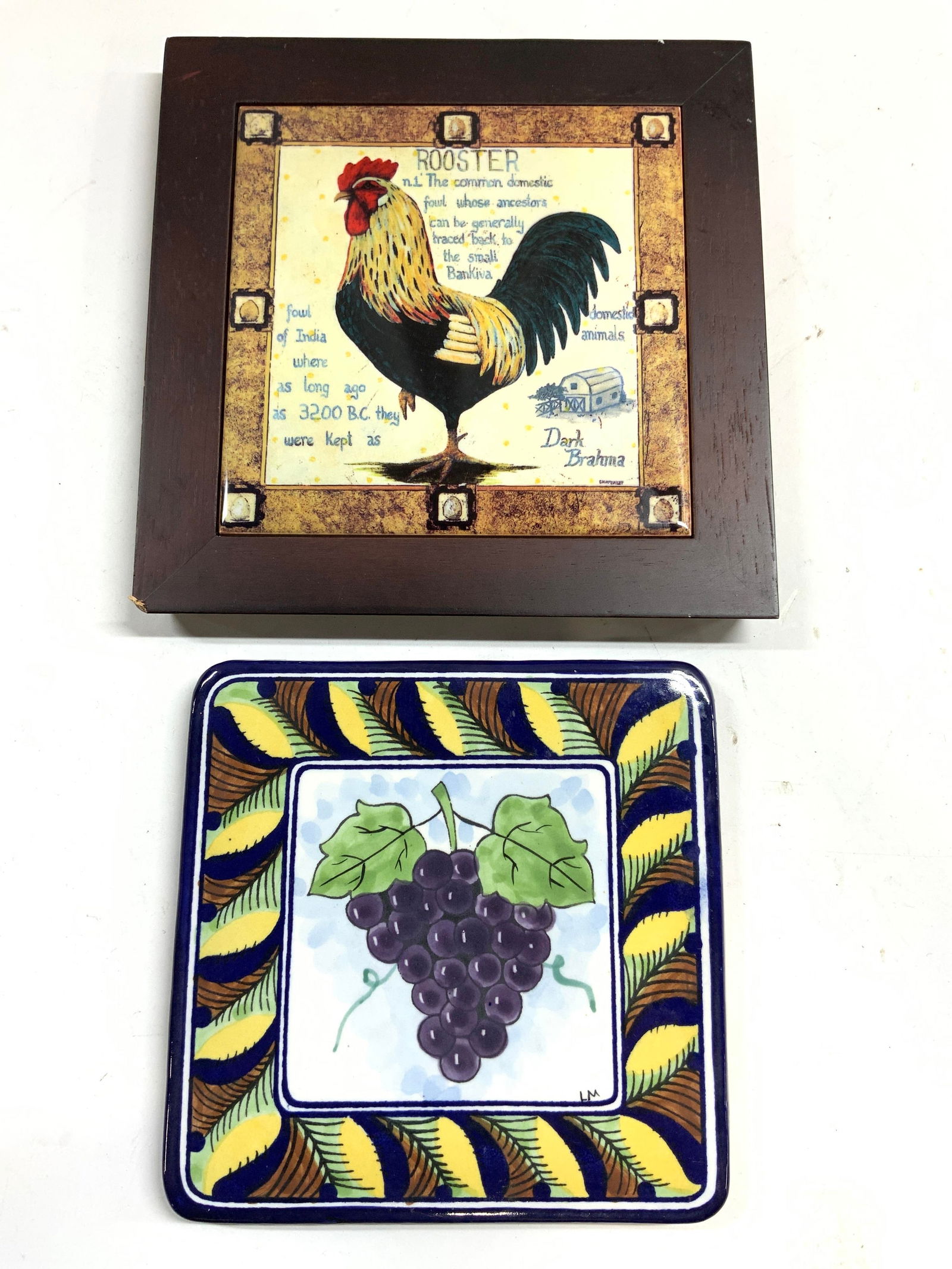 Lot 2 Assorted Porcelain & Wood Trivets: Lot includes 2 trivets. Lot includes 1 porcelain trivet with wood veneer frame & rooster detail, & 1 Ceramic trivet with multi toned grape and leaf detail. Largest trivet measures approximately 8 inch