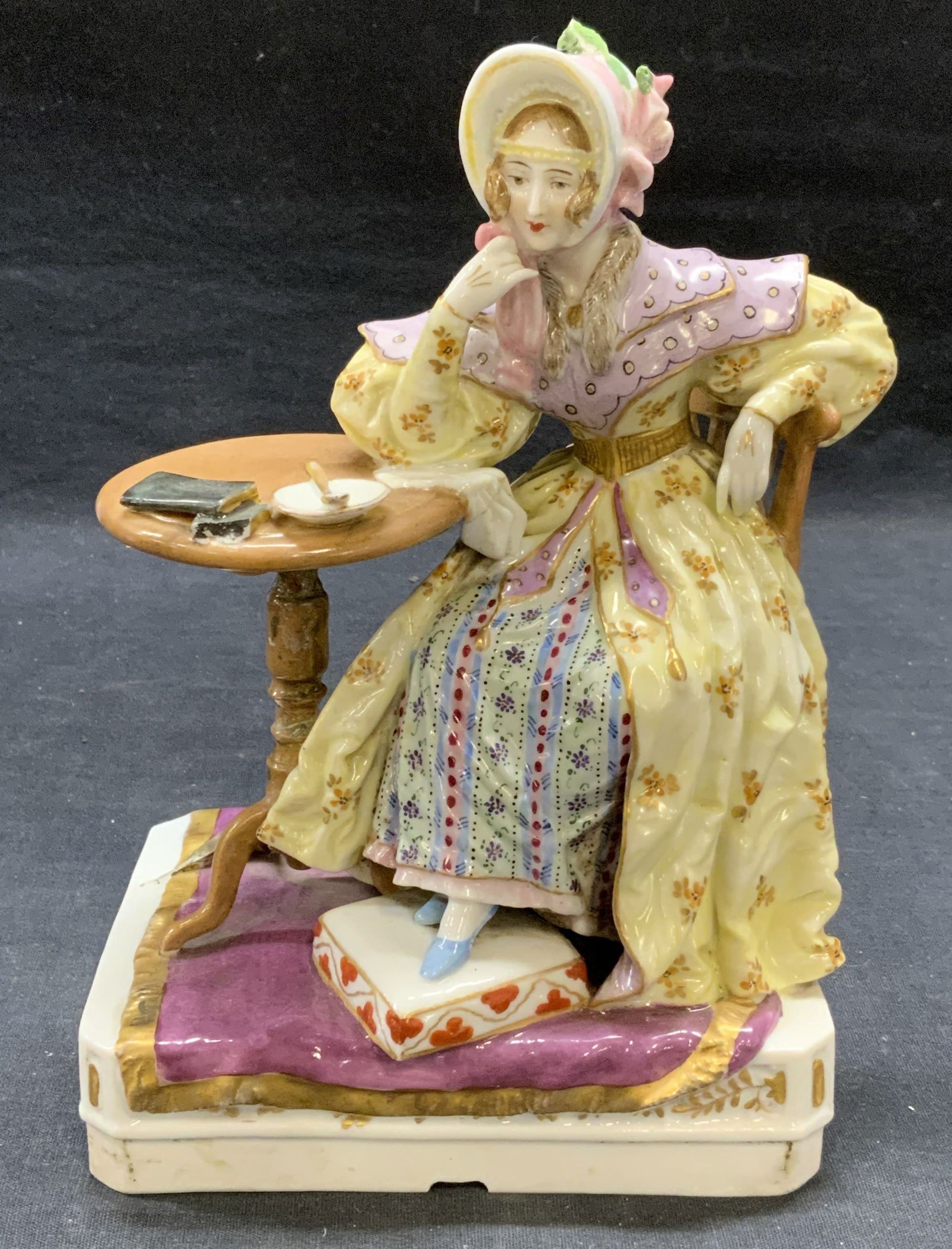 Antique Hallmarked Porcelain Woman Figural: Antique hallmarked porcelain figural. Figure of a woman wear dress and bonnet at table. Hand painted in yellow and purple with gilt decoration. Hallmarked with circle and three dots to underside. Meas