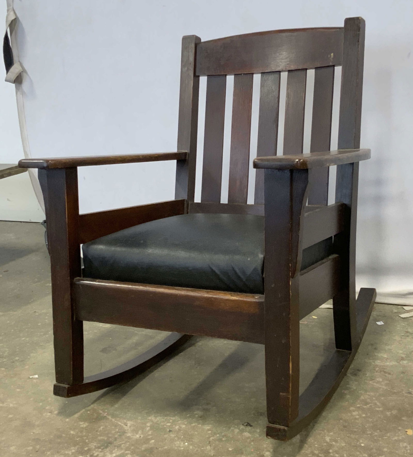 Vintage STICKLEY Style Mission Rocking Chair: A vintage STICKLEY style mission rocking chair with a black leather style upholstered seat. Deep brown hued finish to the wood. Measures approx. 32.5H x 28W x 29D in. Losses to fabric, separation of w