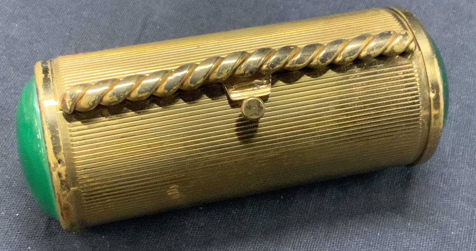 Vntg Gilt Metal Cigarette Case: Vintage cigarette case in a circular shape with horizontal grooves etched within and green resinous caps on each end, with hinged lid and latch. Approx 4 inches long. 1.5 inches wide. Moderate surface