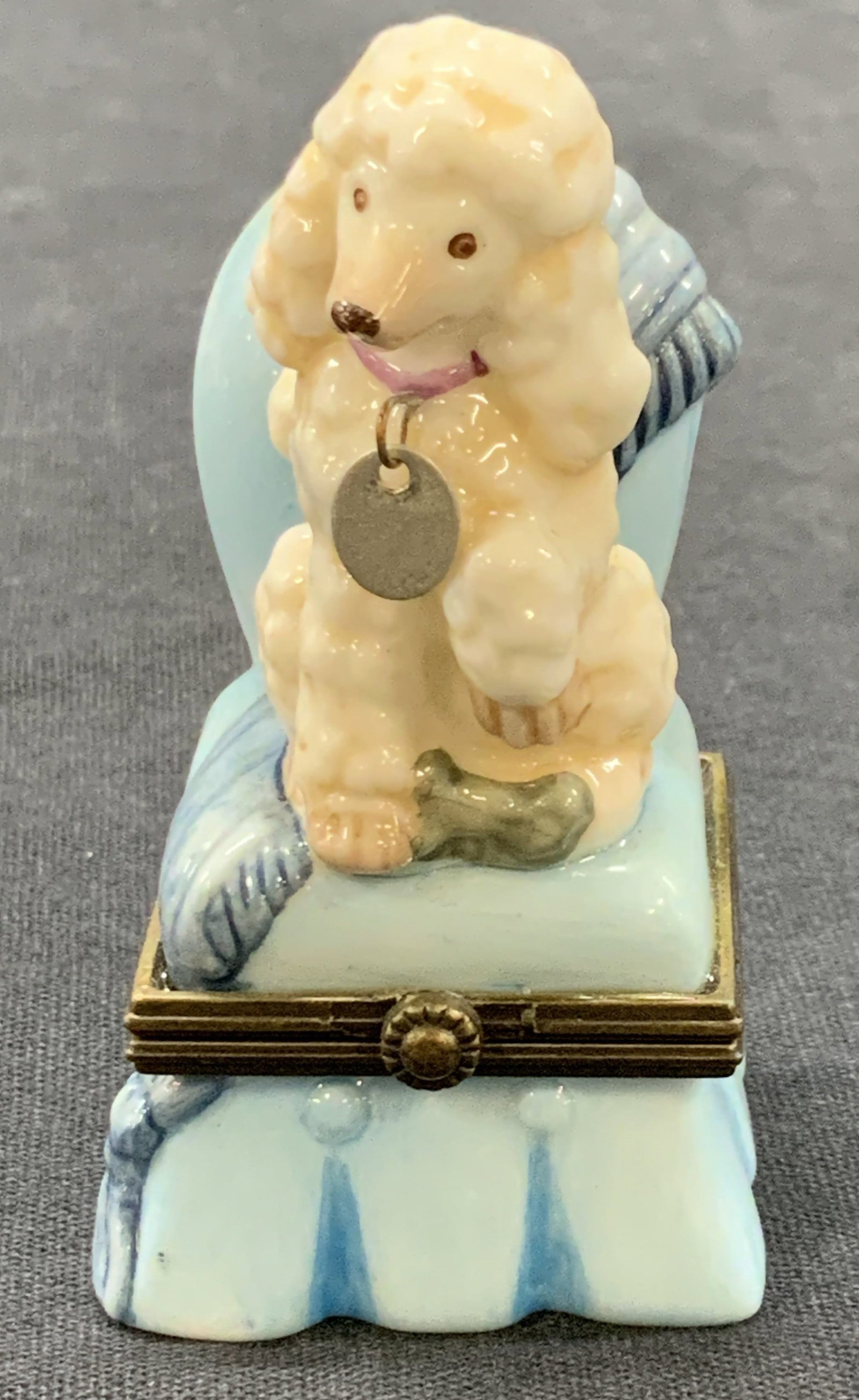 MIDWEST CANNON FALLS Vntg Porc Poodle Pill Box: Vintage Porcelain Poodle in Chair with Dog Tag Trinket Box. Underside reads, Designed by Midwest of Cannon Falls. Poodle is white hued with a purple toned collar. The chair the poodle sits in is a blu