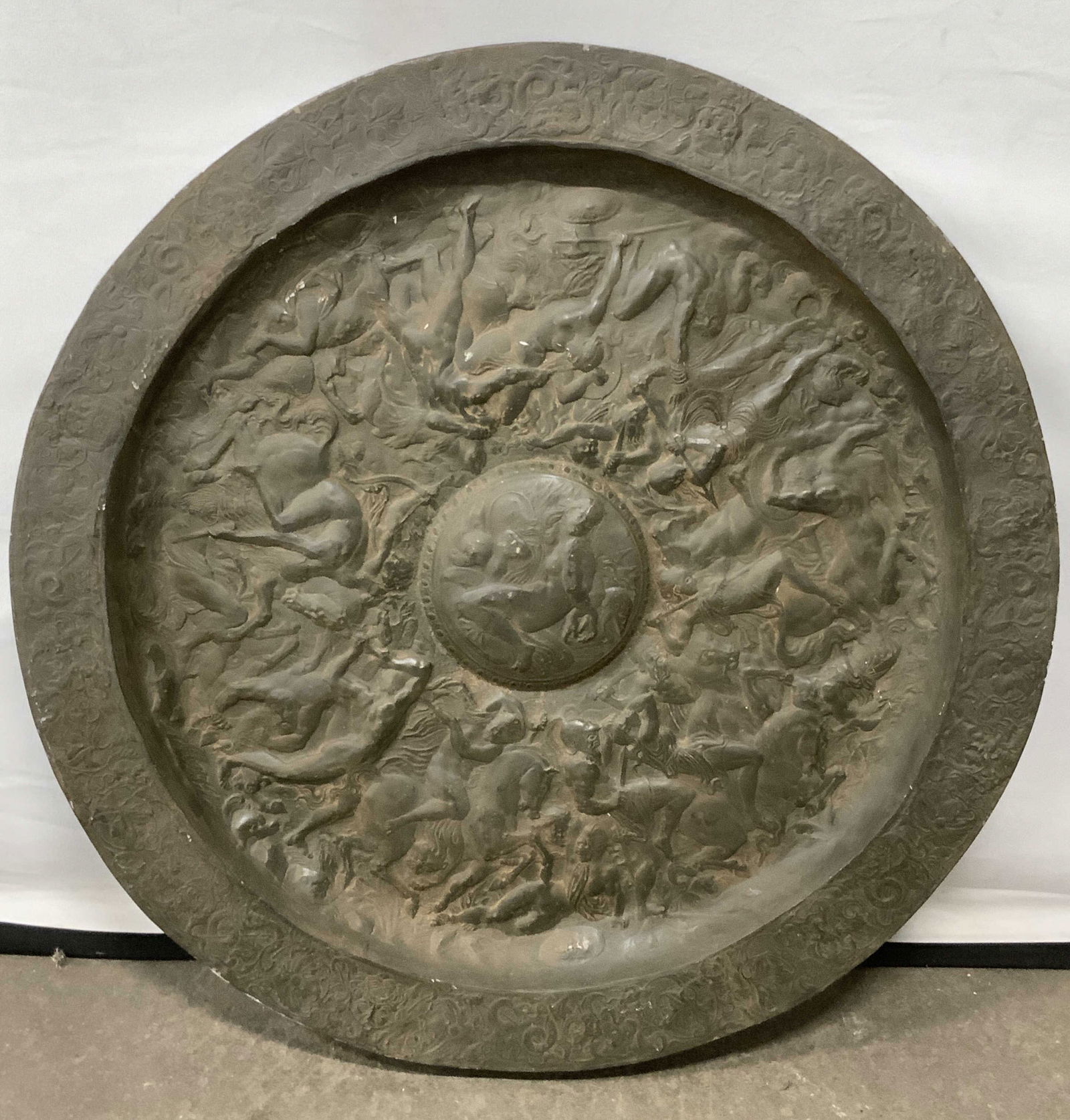 Round Composite Battle Scene Medallion, Wall Art: A round composite battle scene medallion. Dark brown and black hues. Raised scene to obverse features warriors with weapons and on horseback. Center image depicts a seated nude woman. Medallion measur