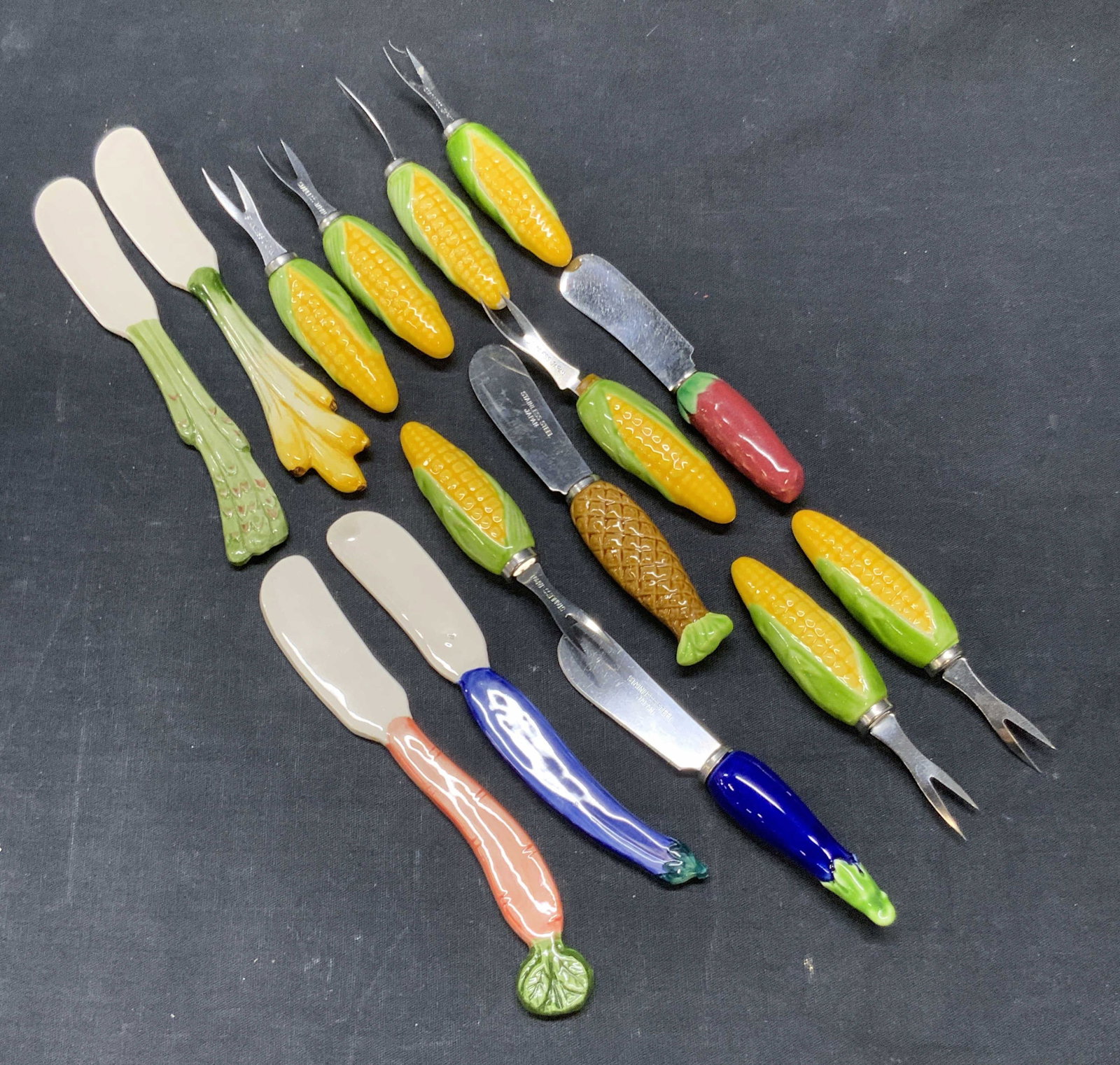 15 Ceramic & Stainless Steel Veg Motif Utensils: A lot consisting of fifteen vegetable / fruit motif utensils. Pieces are crafted from ceramic or stainless steel & ceramic materials. Vegetable motifs include corn, eggplant, carrot, and more. Largest