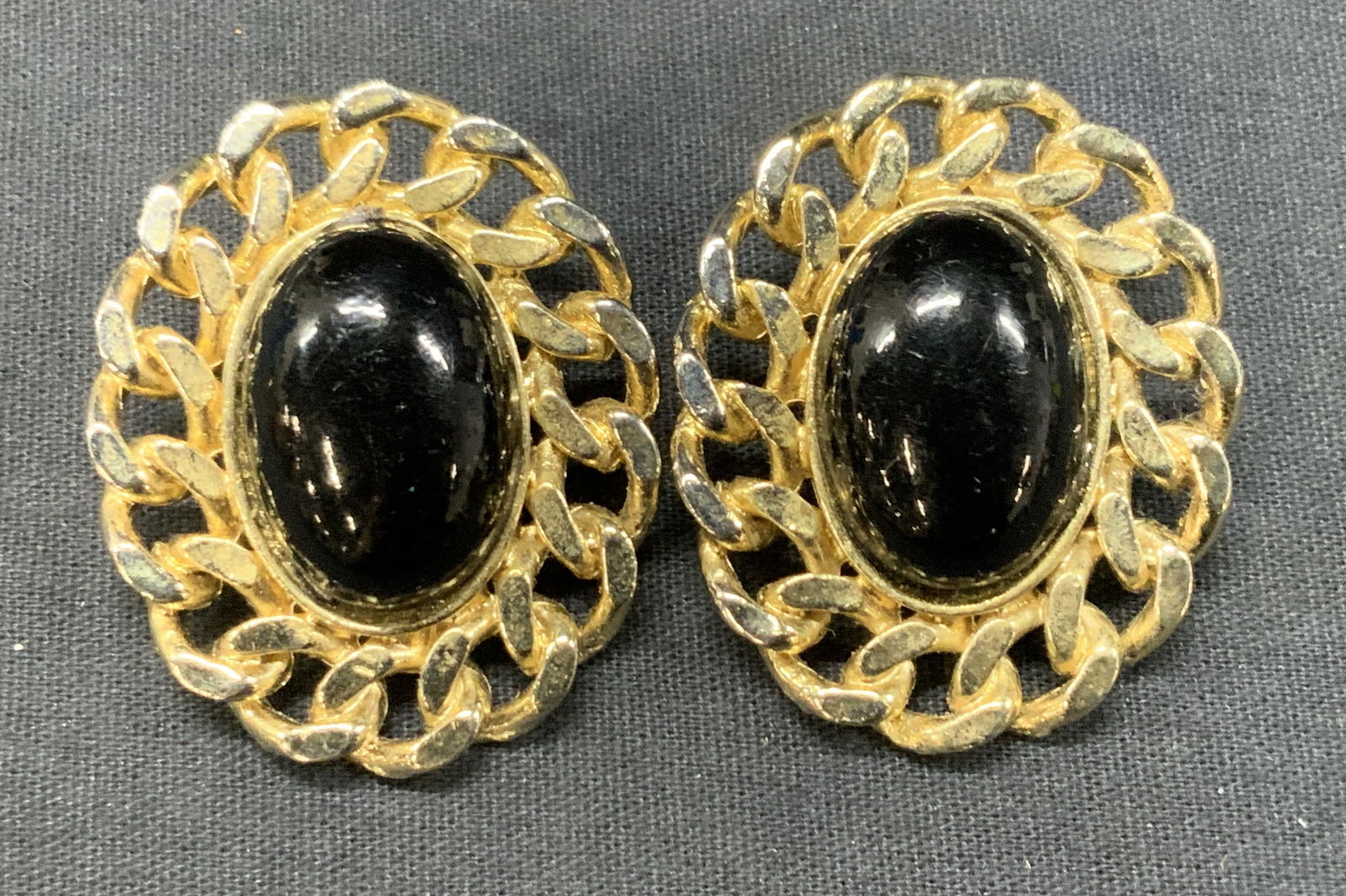 Pr Gold Tn Chain Clip On Earrings: Pair of clip on earrings, gold toned white metal made with chain designs on outer edge and black glass cabochon on center. Approx 2 x 1.75 inches. Moderate losses to gold tone to metal and repairing t