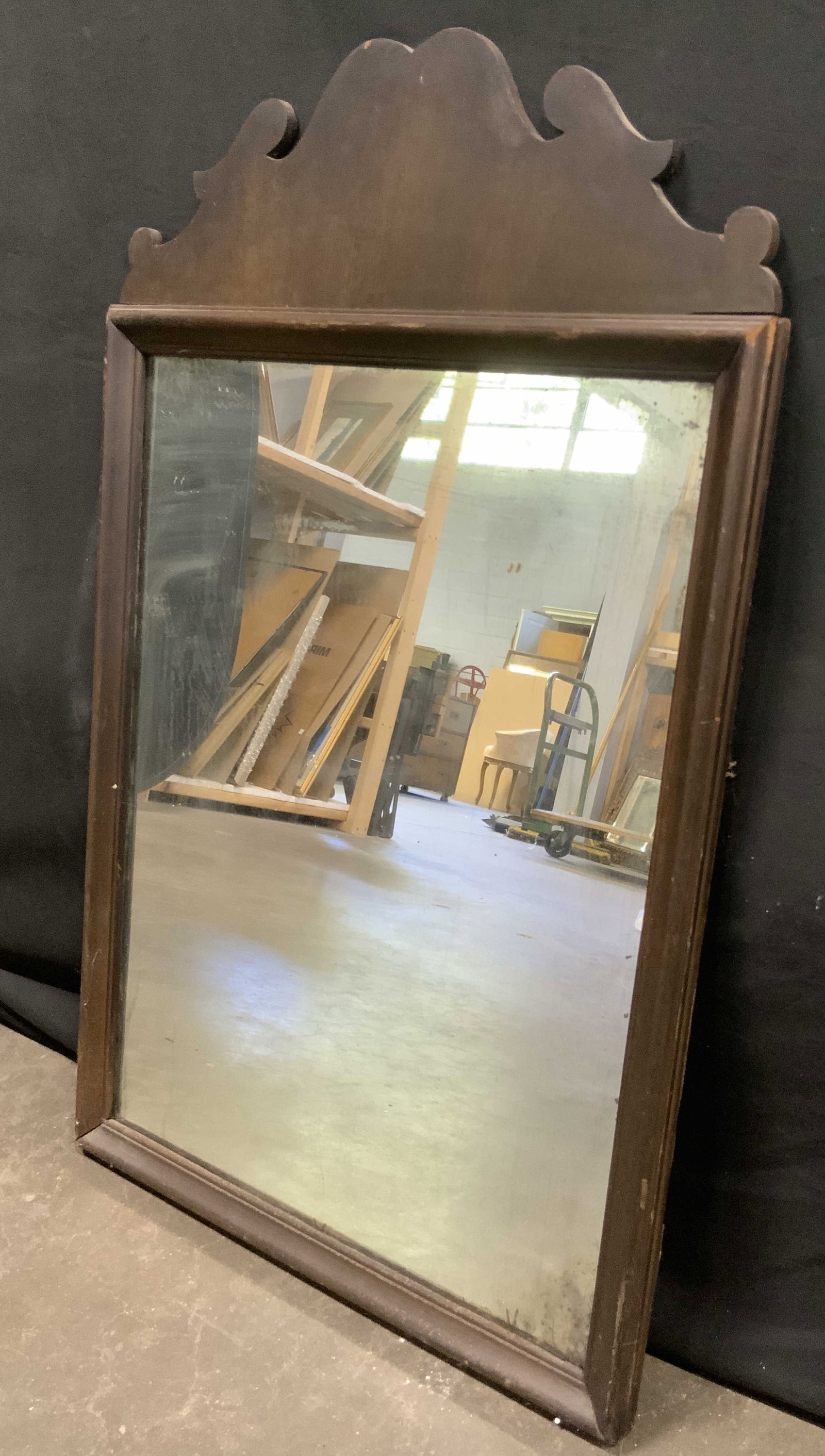 Vintage Wooden Wall Mirror: A vintage wooden mirror with a deep brown hued wood finish. Scrolled detail to the top. Metal hardware to reverse for display. Measures approx. 20W x 33L in. Some discoloration, separation, chipping, 