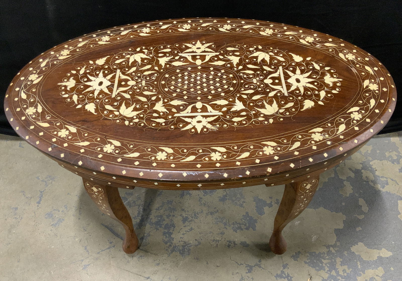 Vintage Inlaid Bone Wooden Side Table: A vintage oval form wooden side table with a bone inlay. Florals, foliage, and geometric patterns to the bone inlay. Curve to the legs. Measures approx. 17H x 18D x 30W in. Some surface wear, discolor