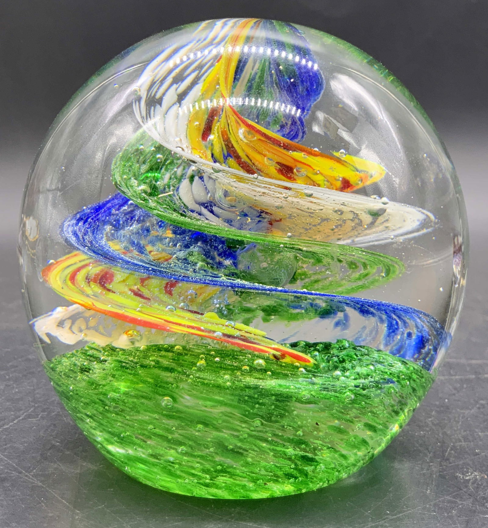 Art Glass Multicolor Swirl Paperweight: Colorless art glass paperweight featuring multicolored swirls to the center. Blue, yellow, green, white, orange, and more colors throughout the swirl. Measures approx. 3.5H x 3.5W in. Measured at wide