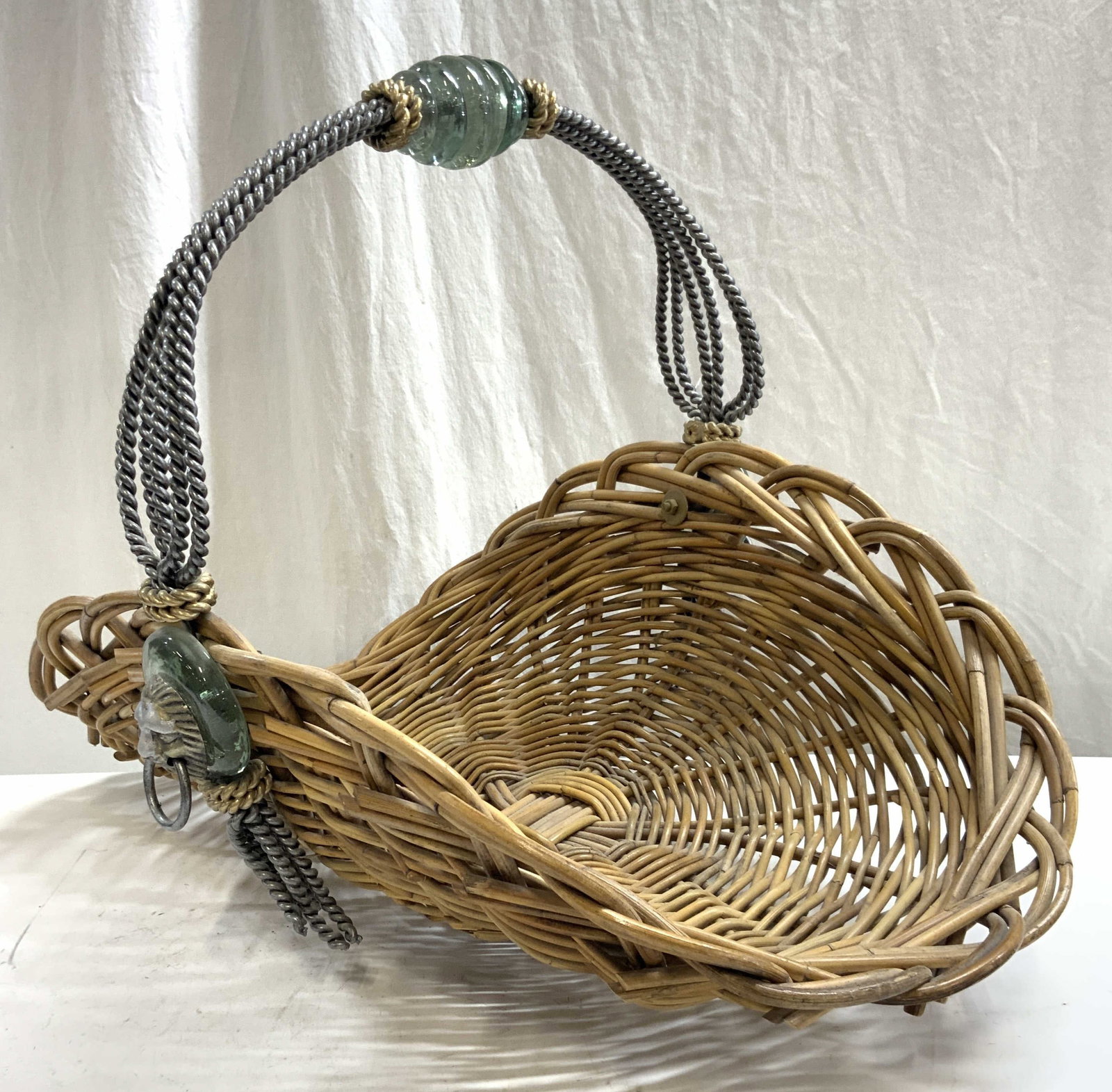 Woven Rattan Firewood Holder W Metal Handle: Firewood holder is woven rattan. Piece is tan toned. Firewood holder has corded metal handle with arch form. Ends of handle have silver toned metal lion head handles. Center of handle appears to be gl