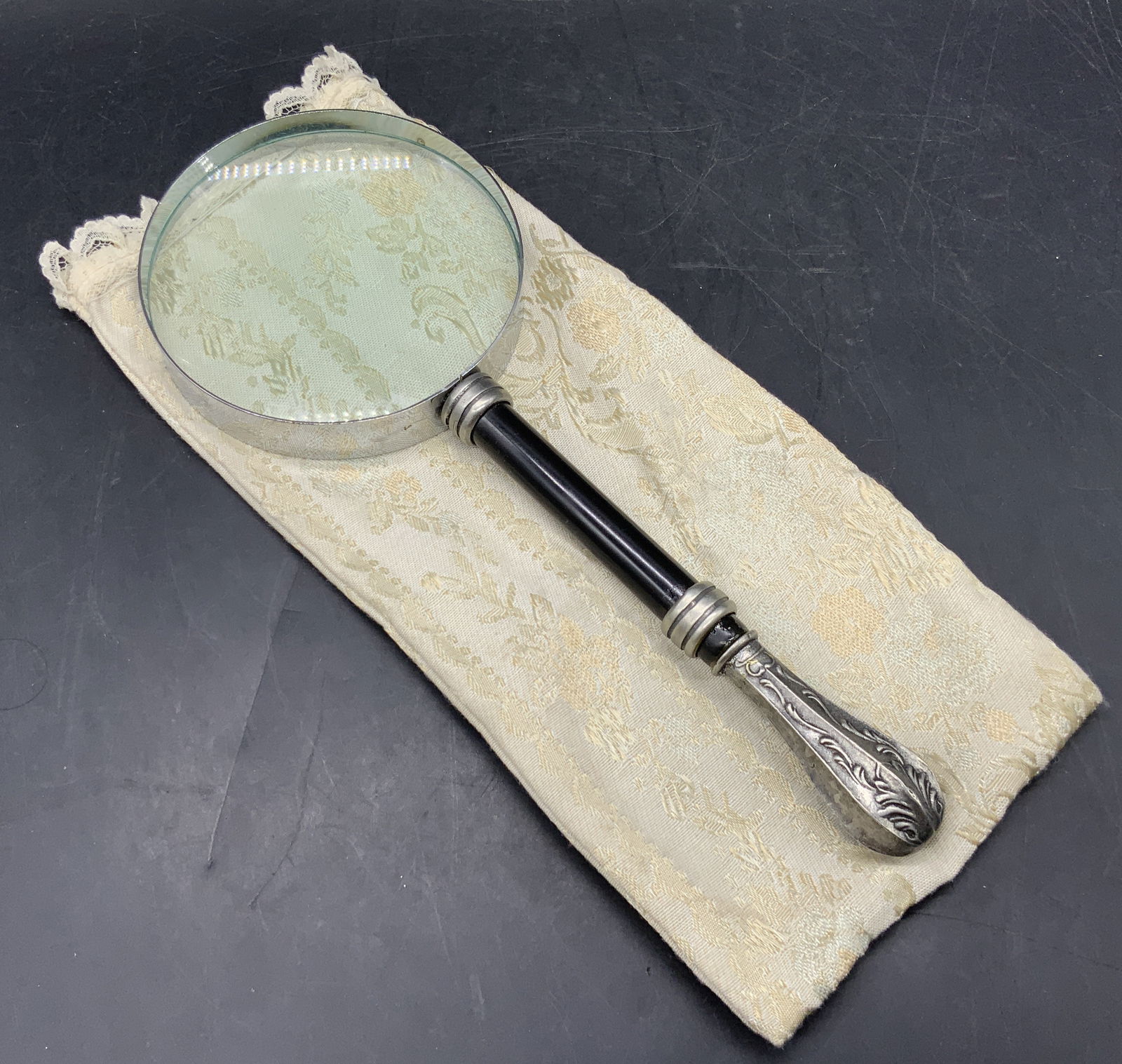 Vintage Magnifying Glass W/ Fabric Case: A vintage magnifying glass featuring a silver toned metal tipped black handle. Magnifying glass measures approx. 10.25L x 4W in. Magnifying glass reads KOREA. Comes in a floral fabric case. Some surfa