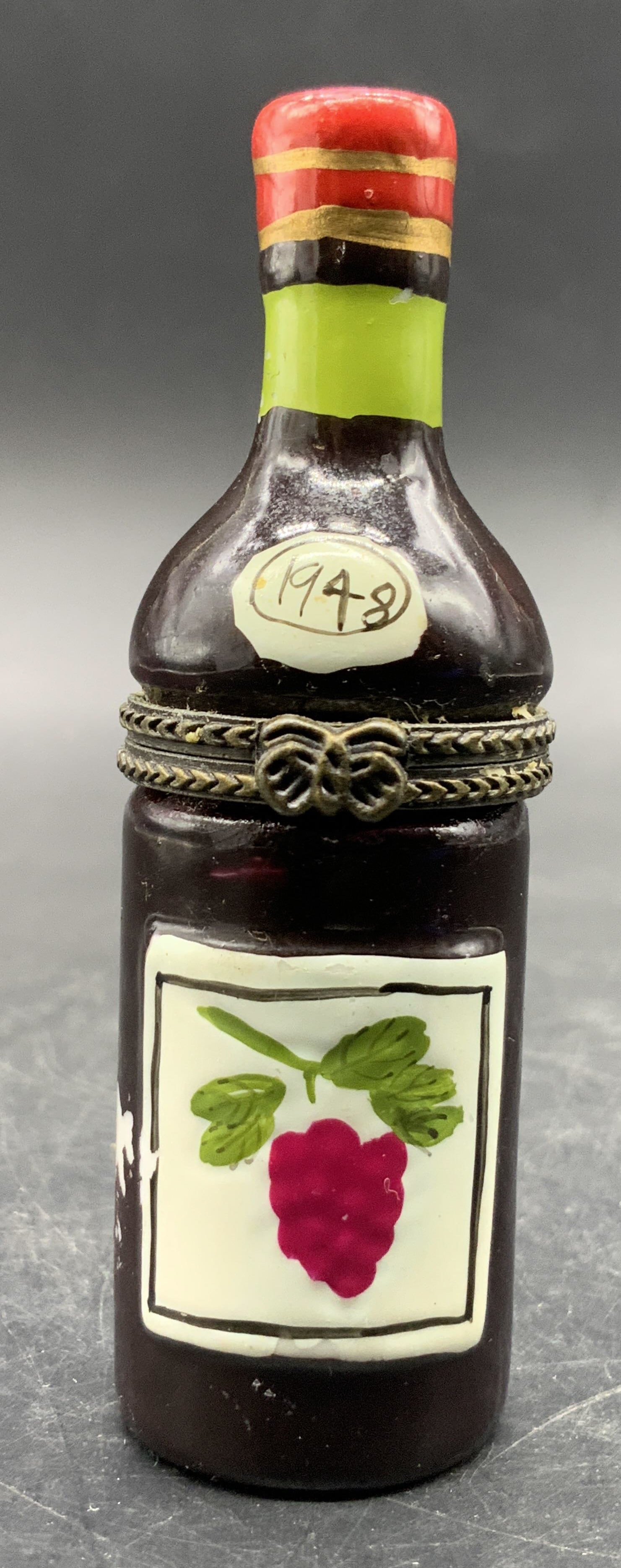 Ceramic Wine Bottle Motif Pill Box: A ceramic wine bottle motif pill box. Grapevine design to the obverse alongside the date 1948. Metal hinge hardware features a ribbon motif to the obverse. Measures approx. 3.25H x 1W in. Losses to pa