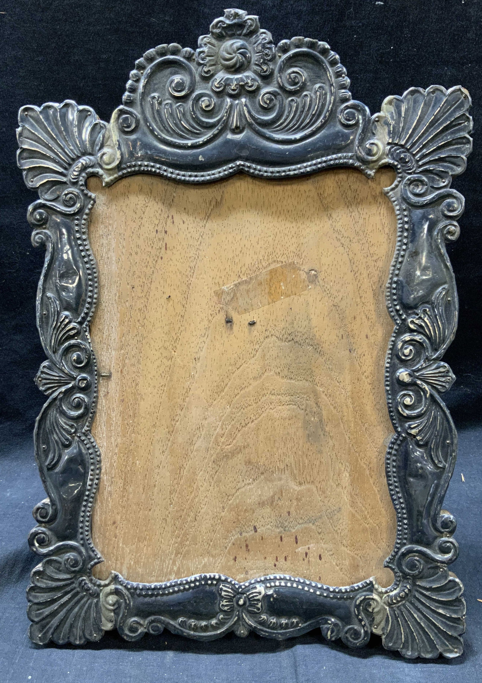 Metallic Ornate Table Top Picture Frame: Wooden frame with thin metallic molding on front. Molding has scrolling motifs. Frame measures approx. 15 H x 10.5 W in. Window measures approx. 9.5 H x 6.5 W in. Backing has hook to close, no loop to