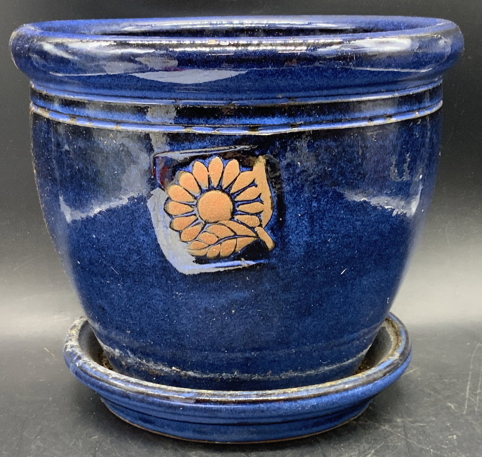 Vintage Blue Glazed Floral Motif Pottery Planter: A vintage blue glazed pottery planter with a floral motif. Thick walled and weighty. Affixed to drainage plate. Measures approx. 7.75H x 8.75W in. Discoloration and surface wear present. Detritus to i