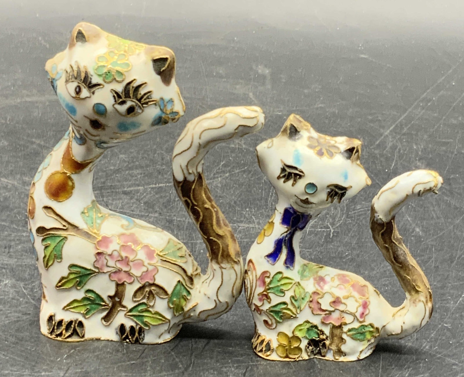 Set 2 Vintage Cloisonne Cat Figurals: A set consisting of two vintage cloisonne cat figurals. Both felines are primarily white with multicolored floral and foliage motifs throughout. Larger cat measures approx. 3.5H x 2.5W in. Some surfac