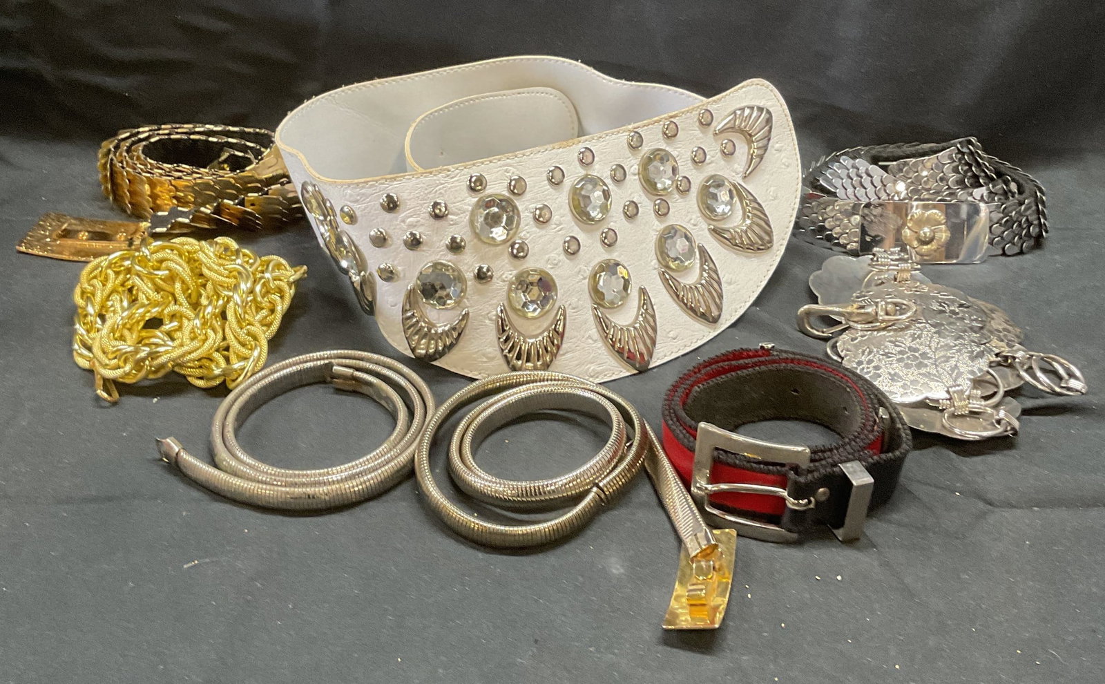 Lot 8 Chain Fashion Belts +: Lot of 8 fashion belts. Includes silver and gold tone chain belts, floral medallion belt, red faux leather belt, and white faux leather and crystal decorated statement belt. Longest belt measures appr