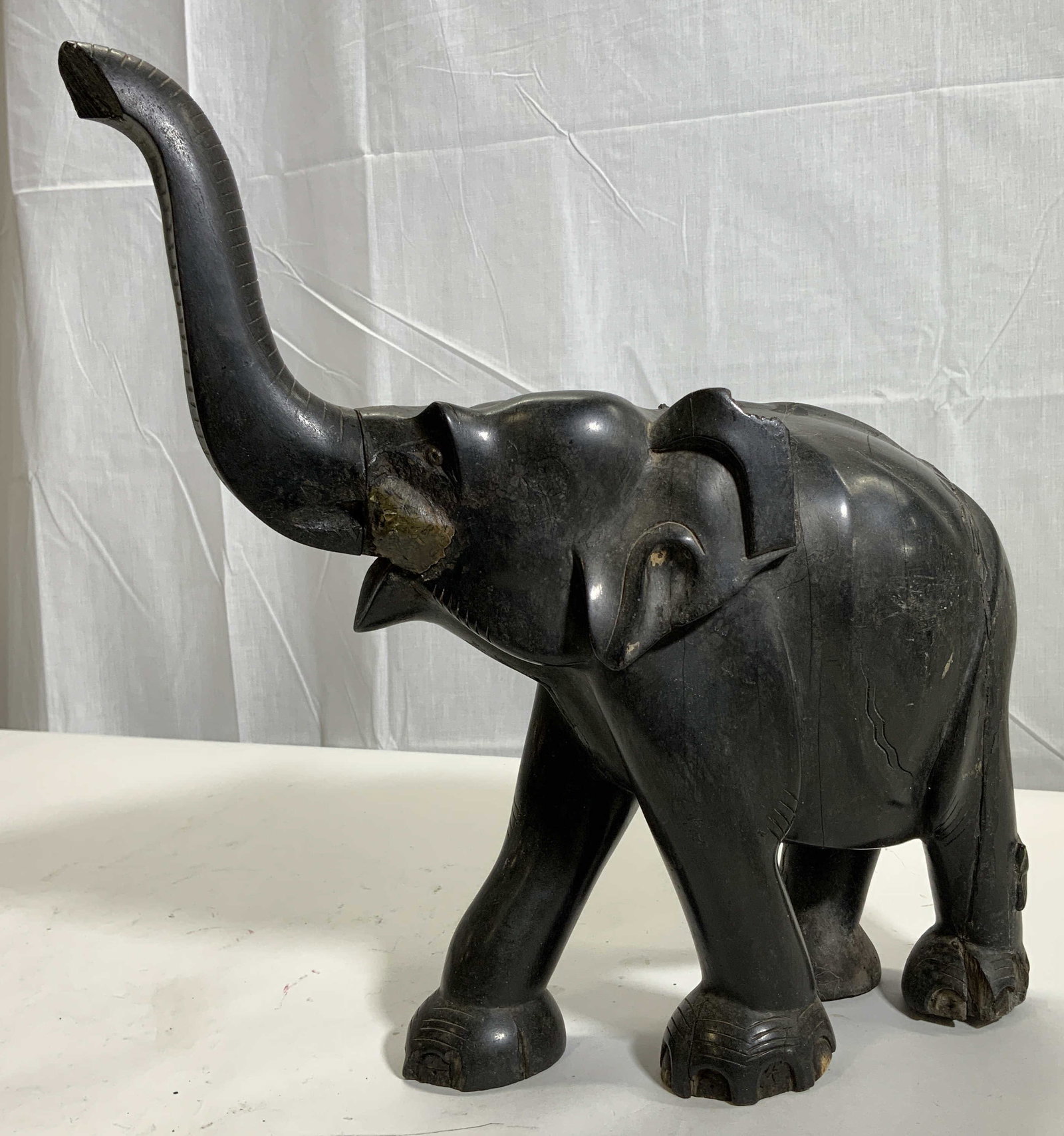 Vintage Carved Black Wooden Elephant Statue: Wooden statue. Subject depicts an elephant walking with his trunk in the air. Statue measures approx. 13 H x 13 W x 4 D in. Piece is heavy set. Trunk is screwed in with screw. Cracks, losses and scrat