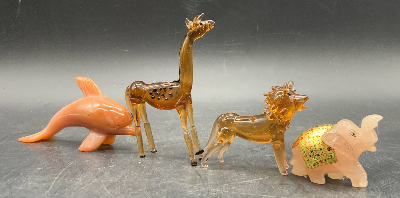 Lot 4 Natural Stone & Glass Animal Figurals: Two stone and two glass animals. The two amber toned glass animals are a lion and a giraffe. The two stone animals are a dolphin and elephant. The elephant has a colorful painted saddle. Giraffe measu
