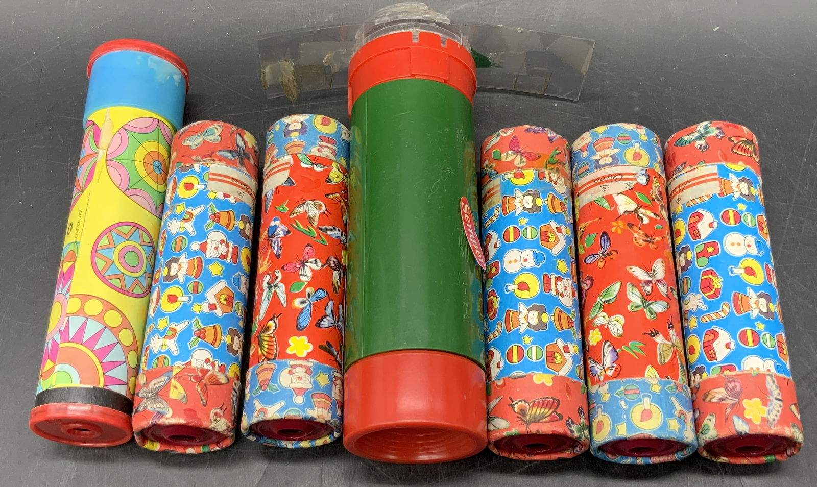 Lot 7 Vintage Kaleidoscope, SEMPER, HK & More: Seven vintage multicolored kaleidoscopes. Patterns include insects, holiday motifs, and geometric shapes. Largest kaleidoscope possesses a sticker which reads SEMPER and measures approx. 6L x 1.75W in