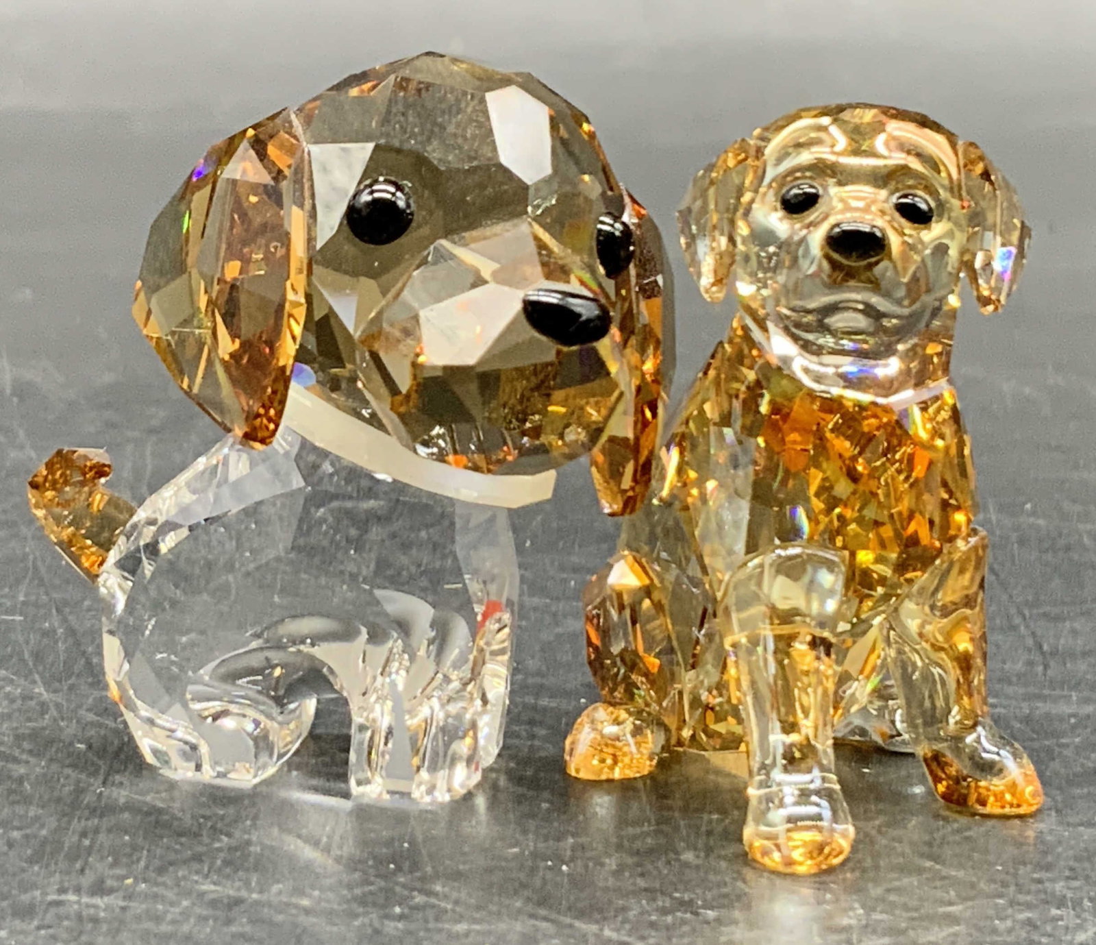 Lot 2 SWAROVSKI Trademarked Crystal Dog Figurals: Two faceted crystal dog figurals. Both pieces are trademarked for SWAROVSKI to the underside. Clear and light brown crystal hues. Larger dog measures approx. 1.5H x 1.25W in. One dog is missing their 