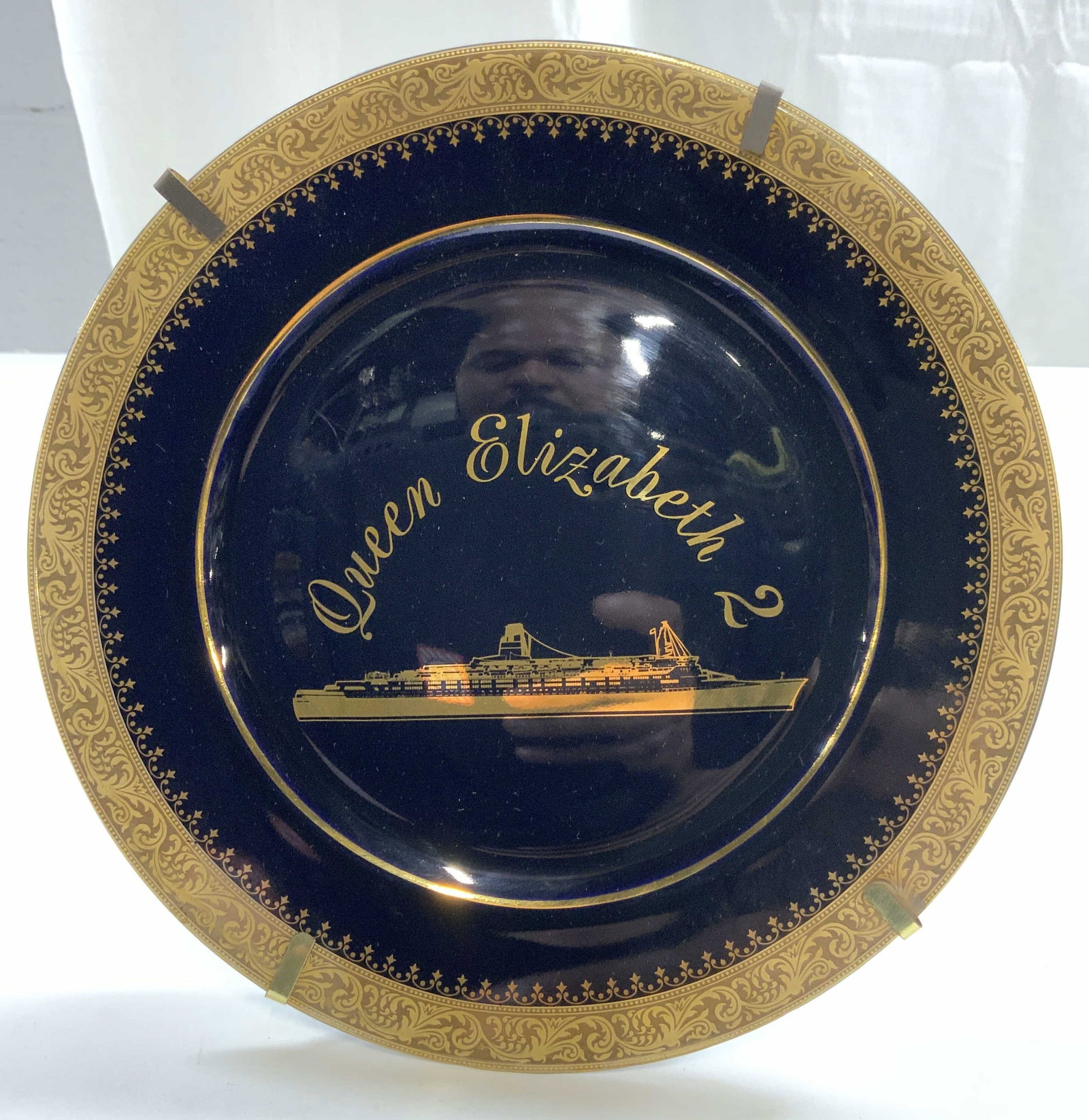 LIMOGES CASTEL Cobalt & Gilt Porc Q Elizabeth Plt: Underside reads LIMOGES CASTEL FRANCE. Plate is cobalt blue tone with ornate gilt detail around rim. Plate sits in holder with stand. Plate reads Queen Elizabeth 2 with image of ship. Piece measures a