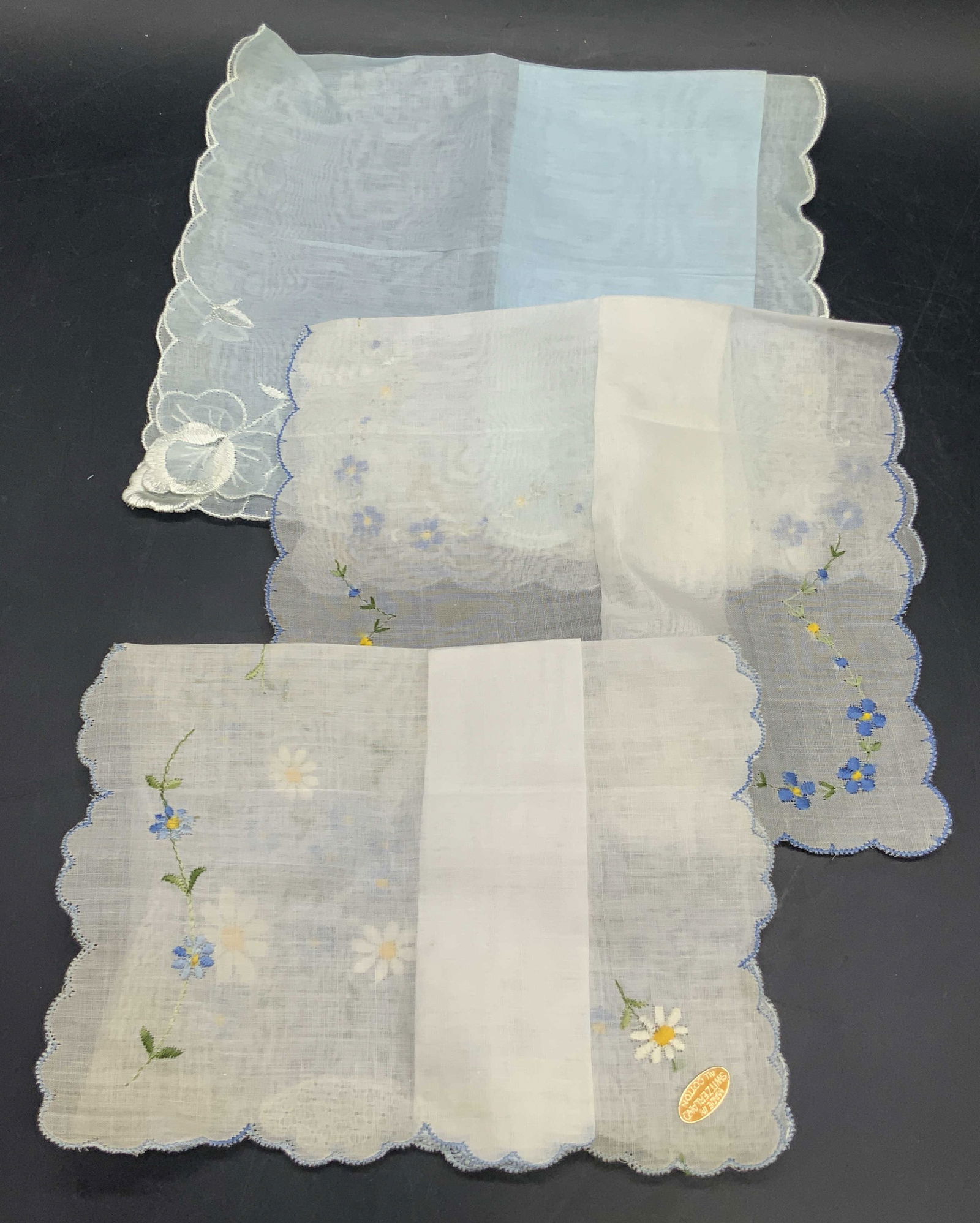 Lot 3 Vntg Floral Embroidered Cotton Handkerchiefs: Three vintage cotton handkerchiefs featuring embroidery. Pieces feature blue forget me nots, daisies, or white floral embroidery. One handkerchief has a sticker to the corner which reads MADE IN SWITZ