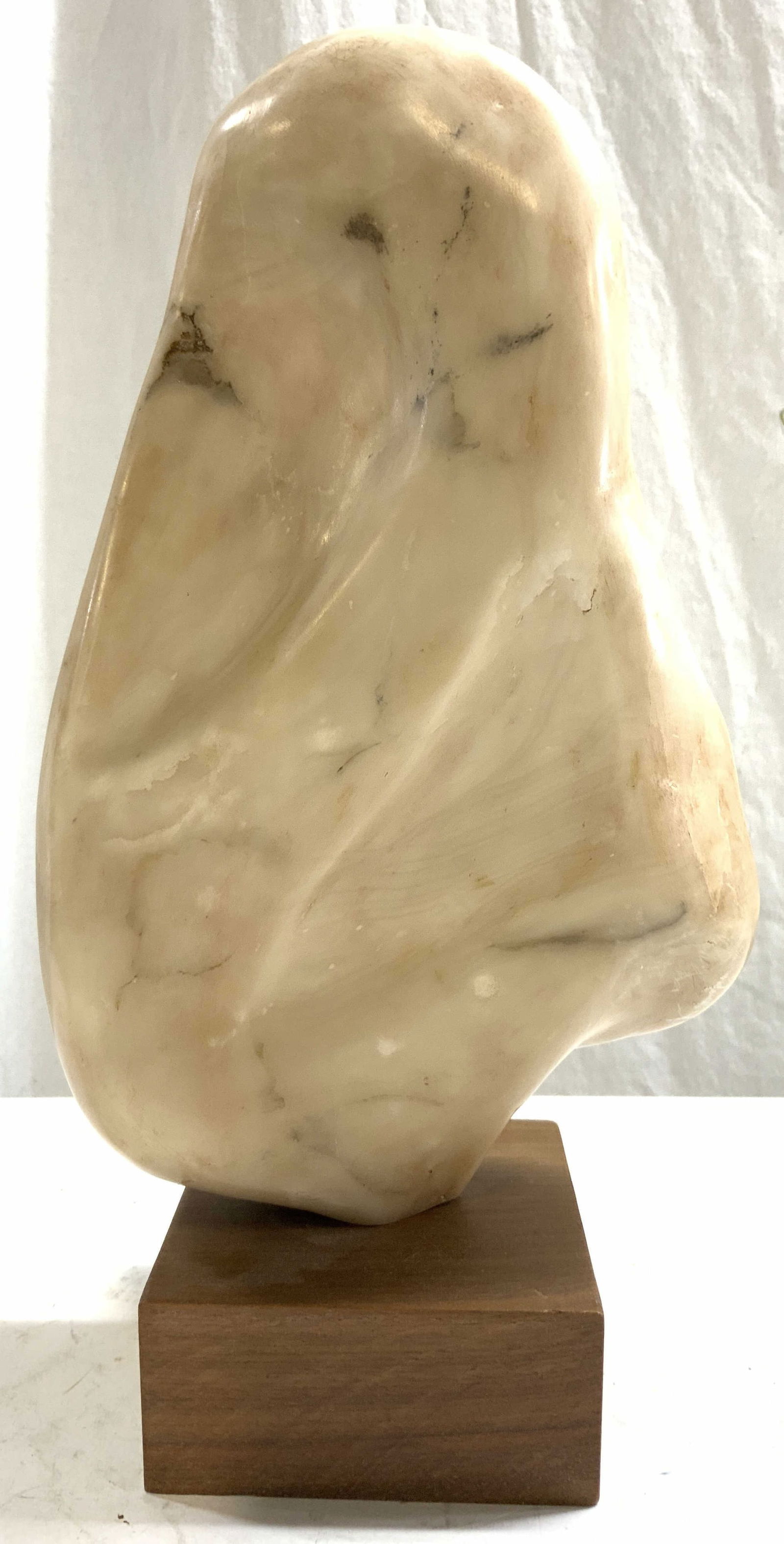 Alabaster Stone Sculpture On Wooden Base: Sculpture is alabaster stone. Piece has various cream and tan tones. Sculpture is attached to square prism wooden base. Piece measures approximately 13.5 inches tall 7.5 inches wide 4 inches deep. Scu